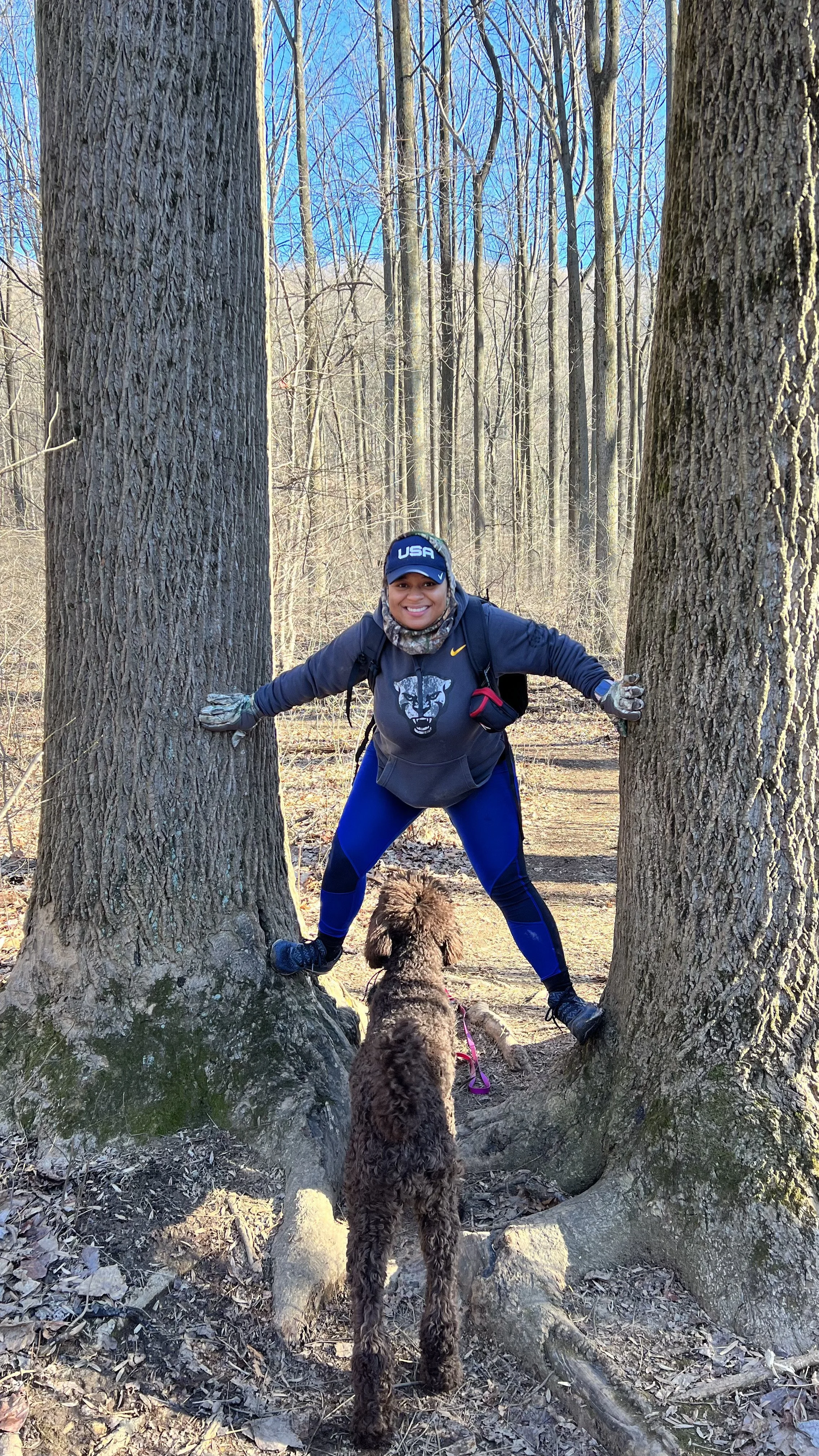 A woman smiling and crouching between two large trees in a forest, wearing athletic clothing, gloves, and a baseball cap that says 'USA.' She is holding onto the trees with her arms extended, and a brown, curly-haired dog is standing with its back to