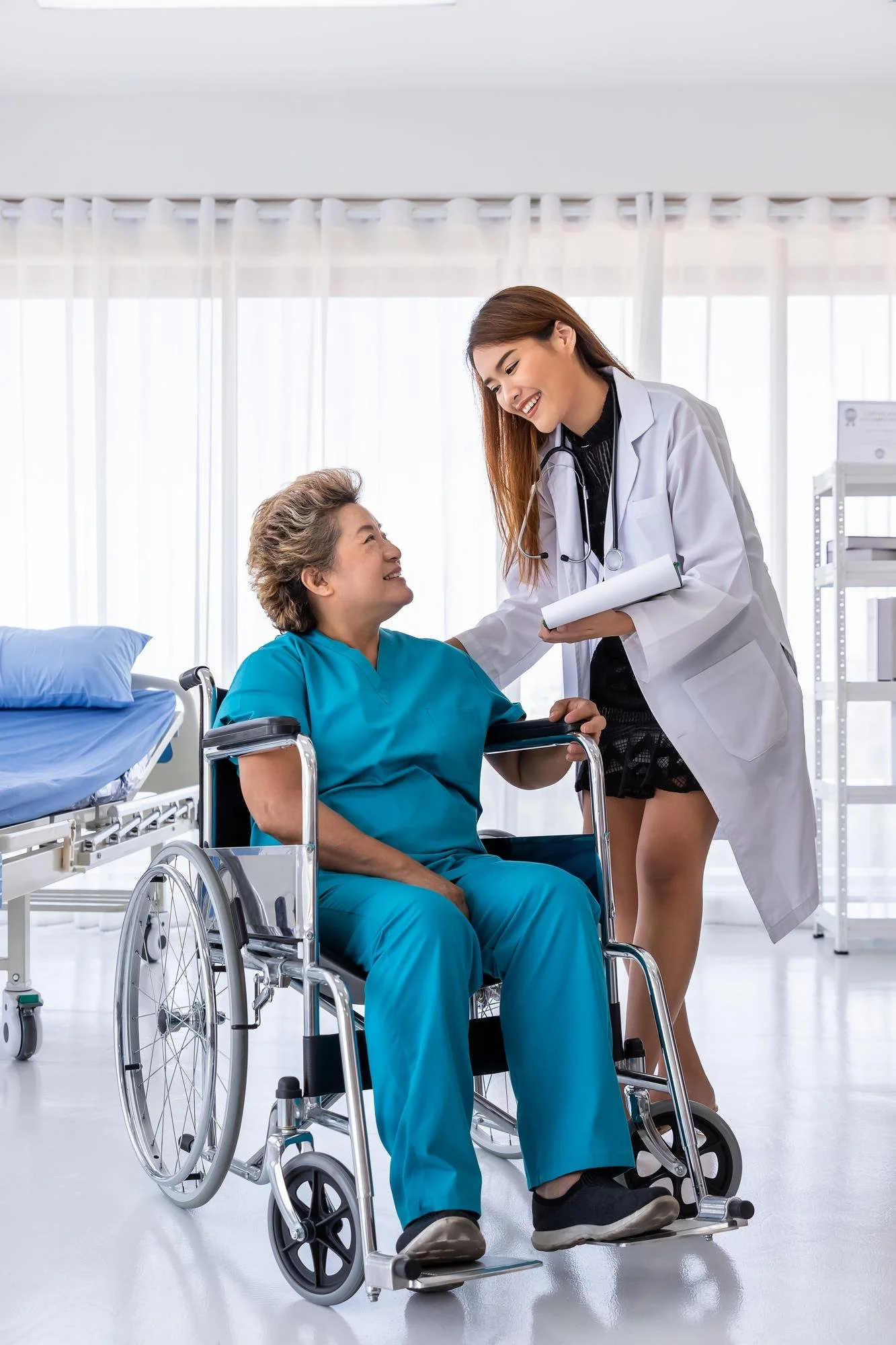 A young female doctor with long hair, wearing a white coat and stethoscope, is smiling and talking to an elderly woman in a wheelchair, who is wearing hospital scrubs, in a bright hospital room.