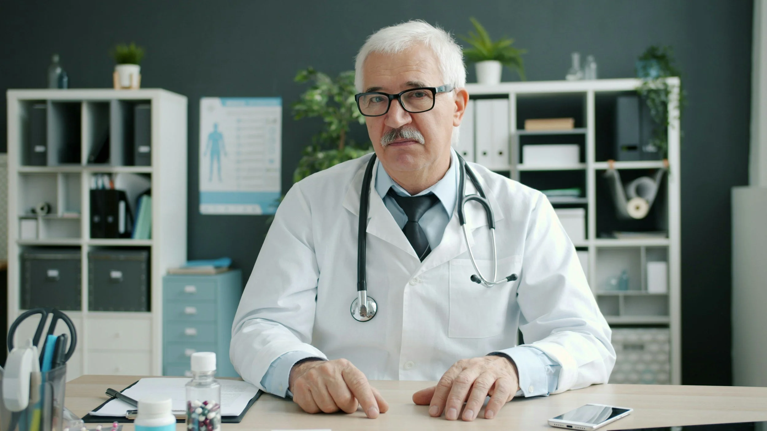 Senior male doctor sitting at a desk in a medical office with medical supplies, a smartphone, and an open notebook in front of him.