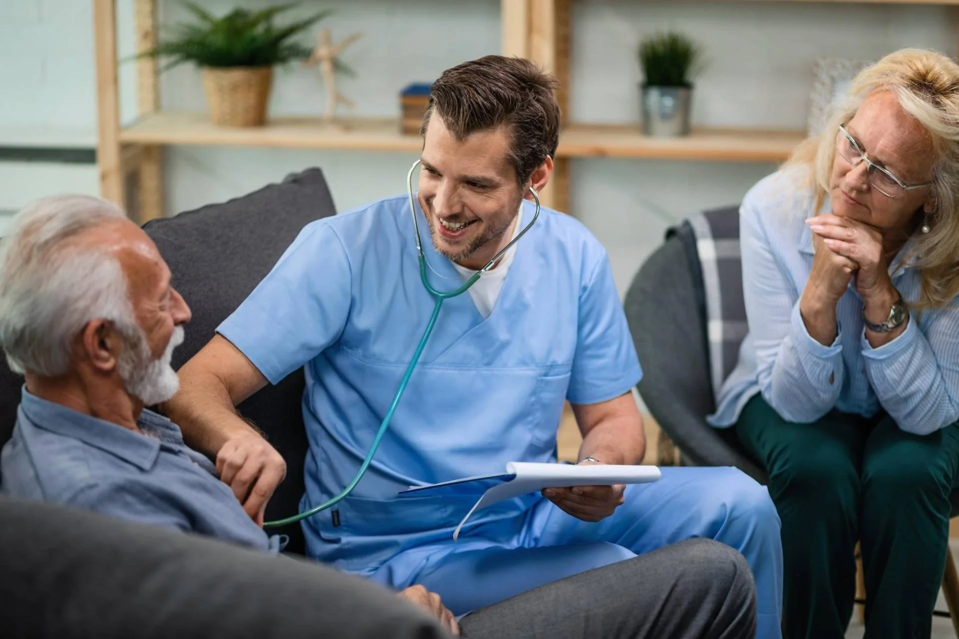 A young male healthcare worker in blue scrubs speaking with an elderly man with gray hair and a white beard, holding a clipboard and wearing a stethoscope around his neck. An older woman with glasses and blonde hair is sitting nearby, listening attentively.