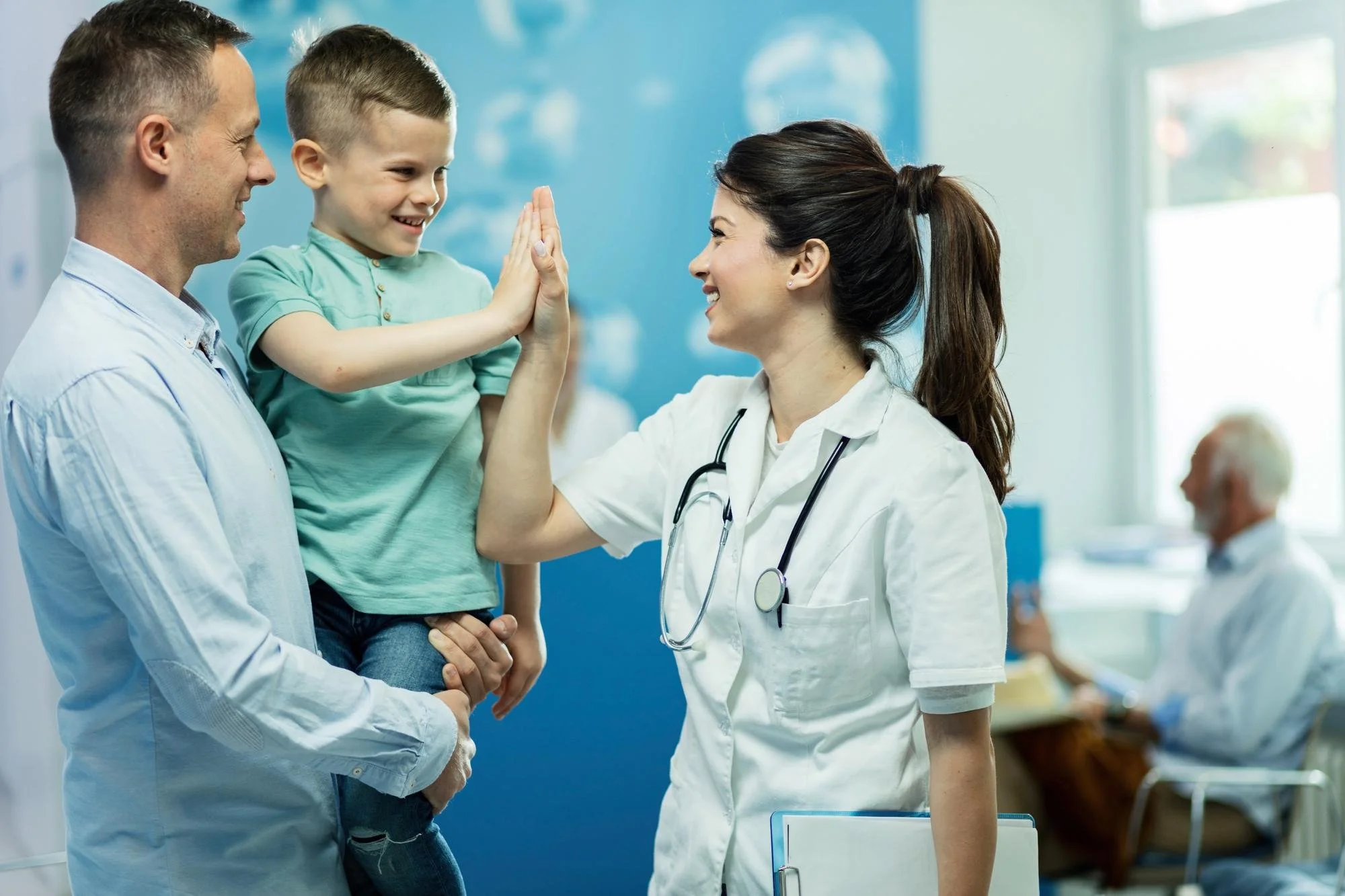 A female doctor giving a high five to a young boy, who is being held by a man, in a medical office. The girl and boy are smiling, and a man in a white coat is in the background.