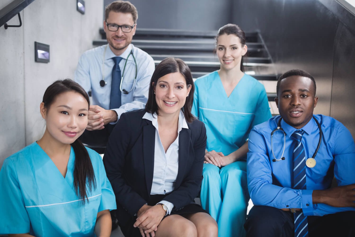 Group of five healthcare professionals, including nurses in scrubs and doctors in formal attire, sitting and standing on stairs in a hospital or medical facility
