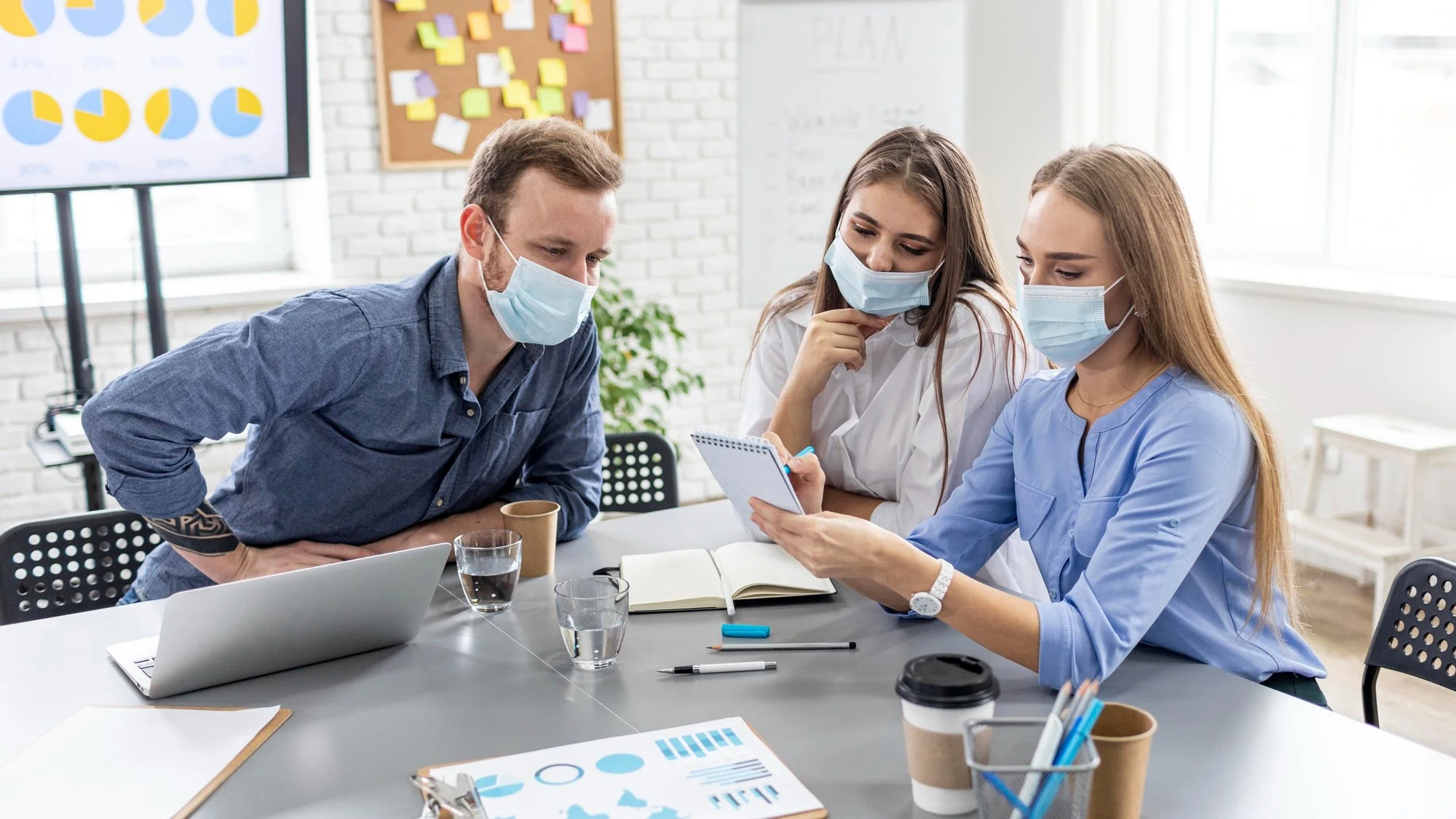 Three people wearing masks sitting at a table in a meeting room, looking at a tablet and discussing. There are notebooks, pens, glasses of water, and coffee cups on the table.
