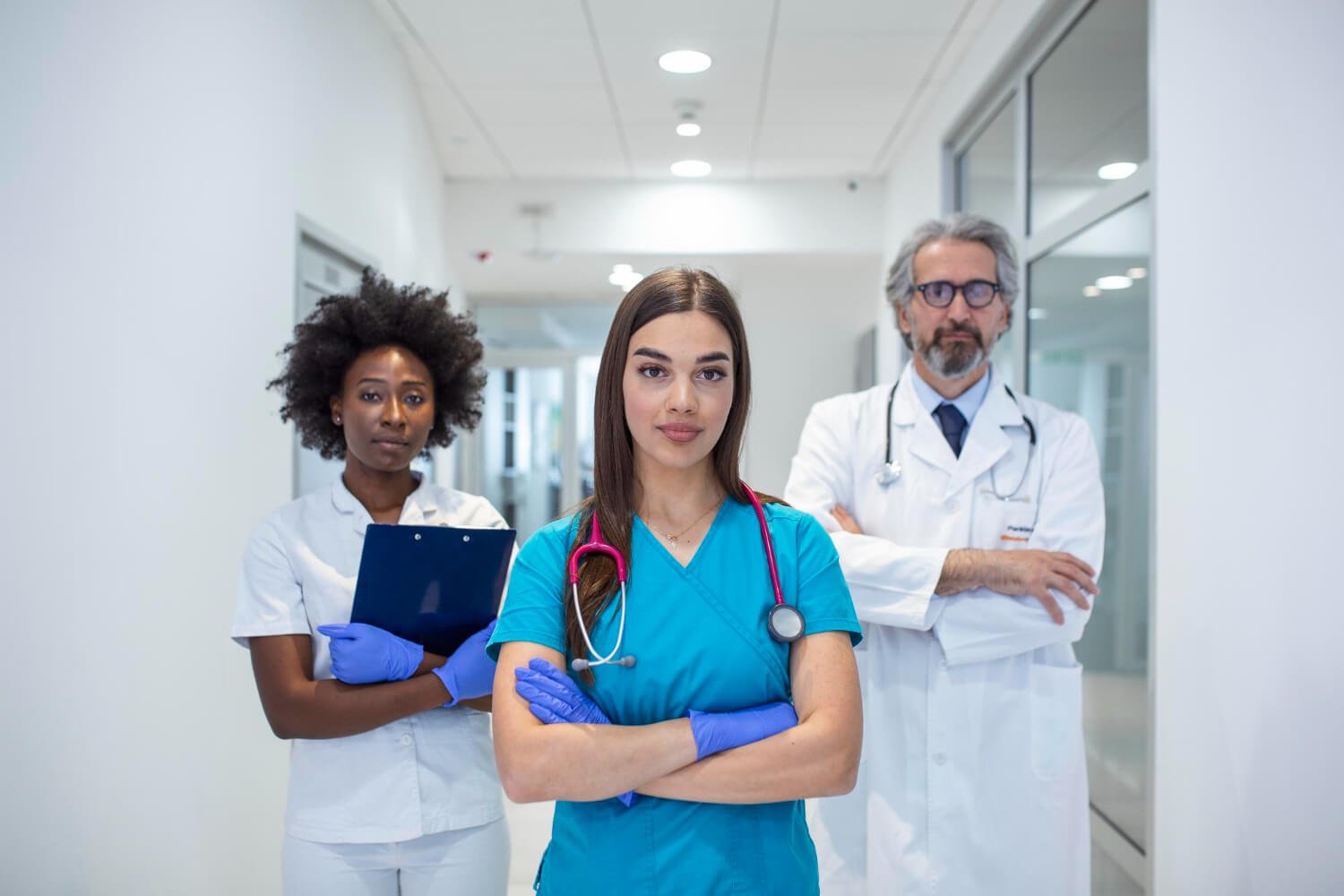 Three healthcare professionals, two women and one man, standing in a hospital corridor. The woman in the center is wearing blue scrubs with a stethoscope around her neck and has her arms crossed. The woman on the left is holding a clipboard and wearing white scrubs and purple gloves. The man on the right, with gray hair and a beard, is wearing white coat and glasses, standing with his arms crossed.
