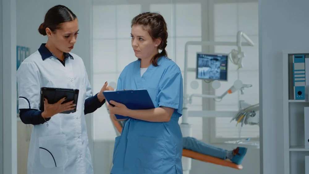 Two female nurses having a discussion in a hospital room, one holding a tablet and the other a clipboard.