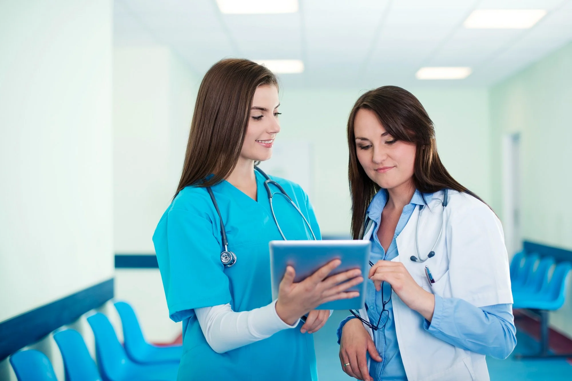 Two female healthcare professionals in scrubs looking at a tablet together in a hospital corridor.