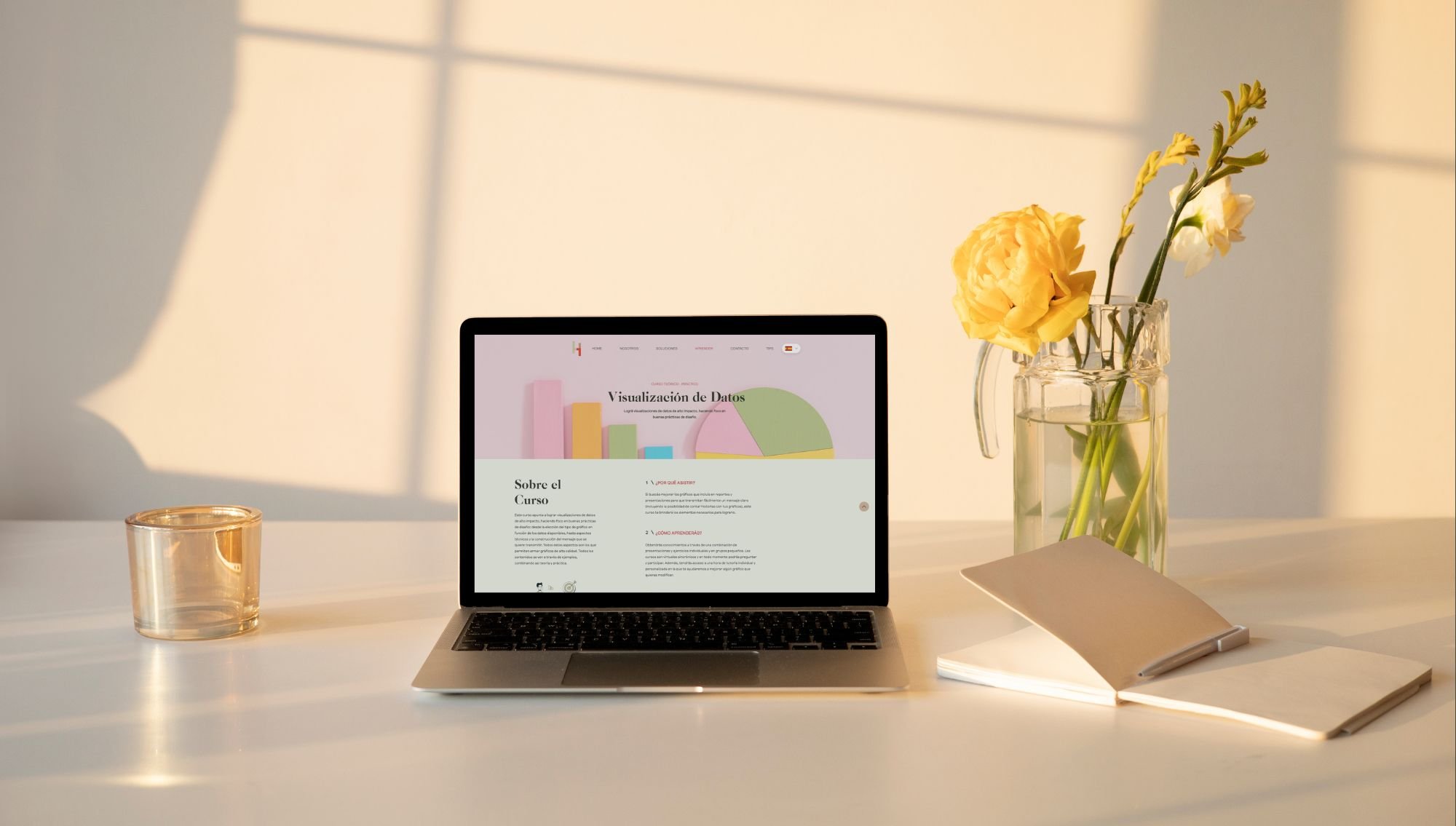 A workspace with a laptop displaying a data visualization webpage in Spanish, an open notebook, a glass of water, a vase with yellow and white flowers, and sunlight casting shadows on a white desk.