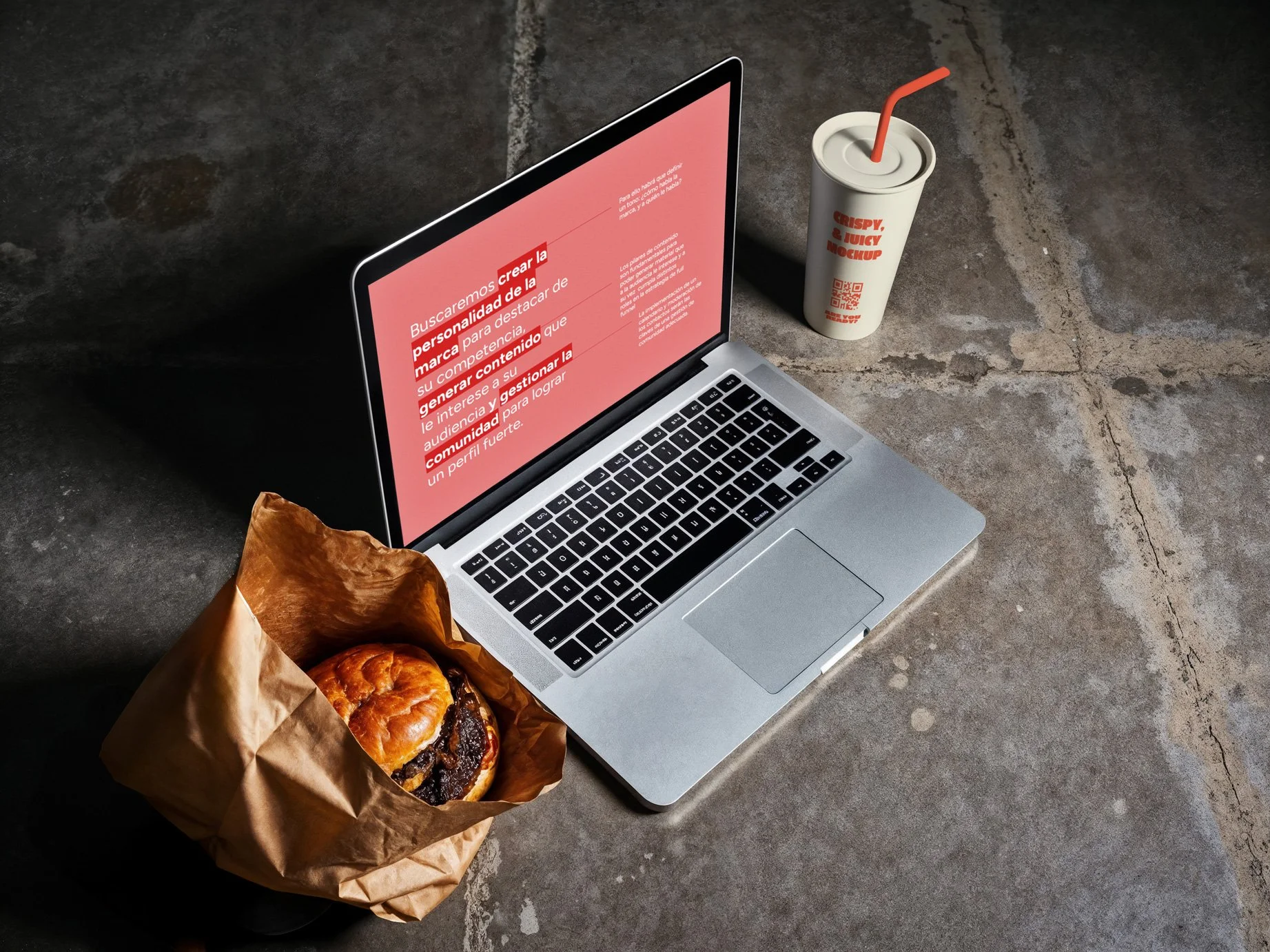 A silver laptop on a concrete floor displaying red text on the screen, a brown paper bag with a croissant sandwich, and a white drink cup with a straw and red text that reads 'CRISPY & JUICY MOCKUP'.