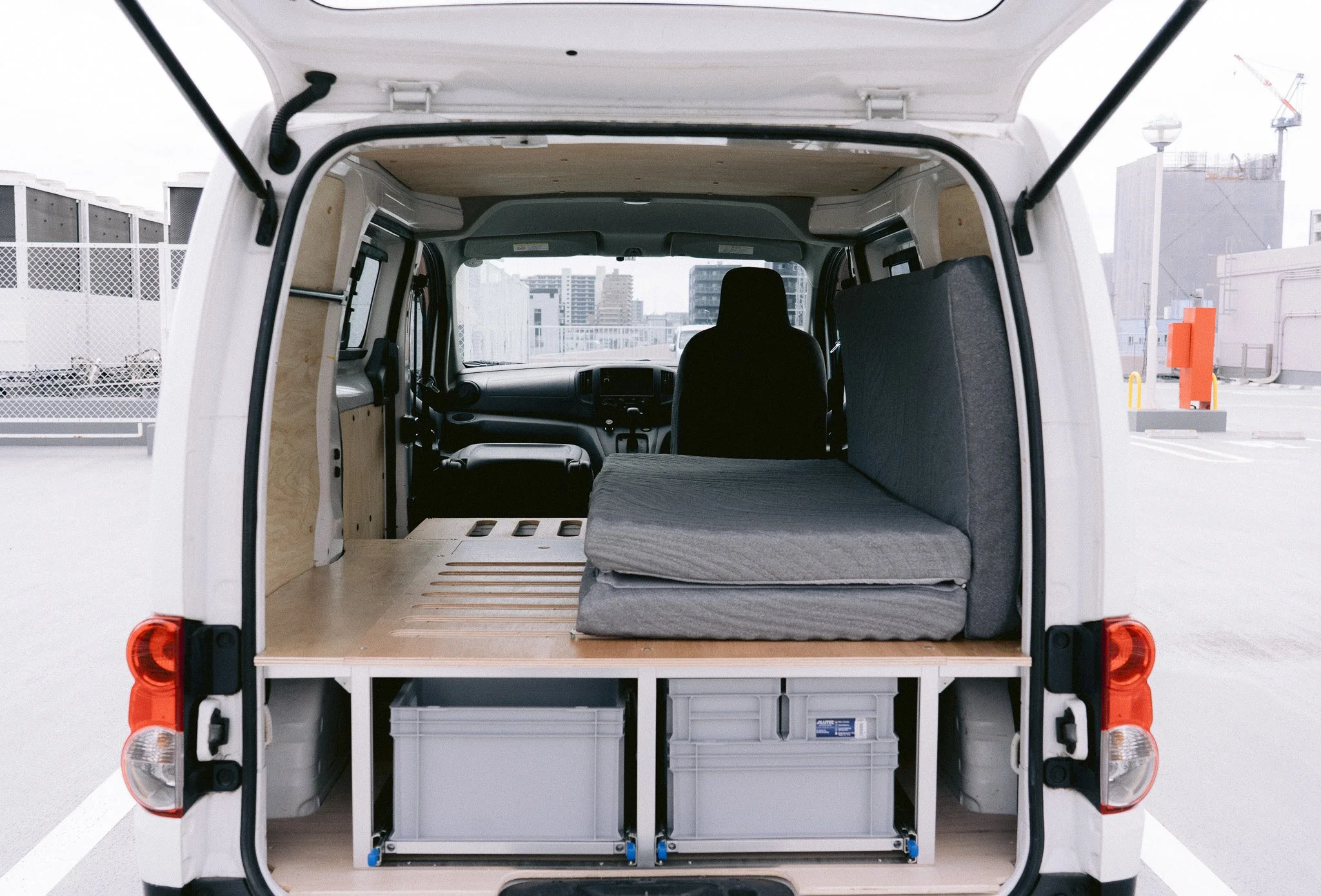 白いバンの後部 interior, 荷台は木製の床と収納ボックスがあり、フラットな睡眠スペースとして使用されている。