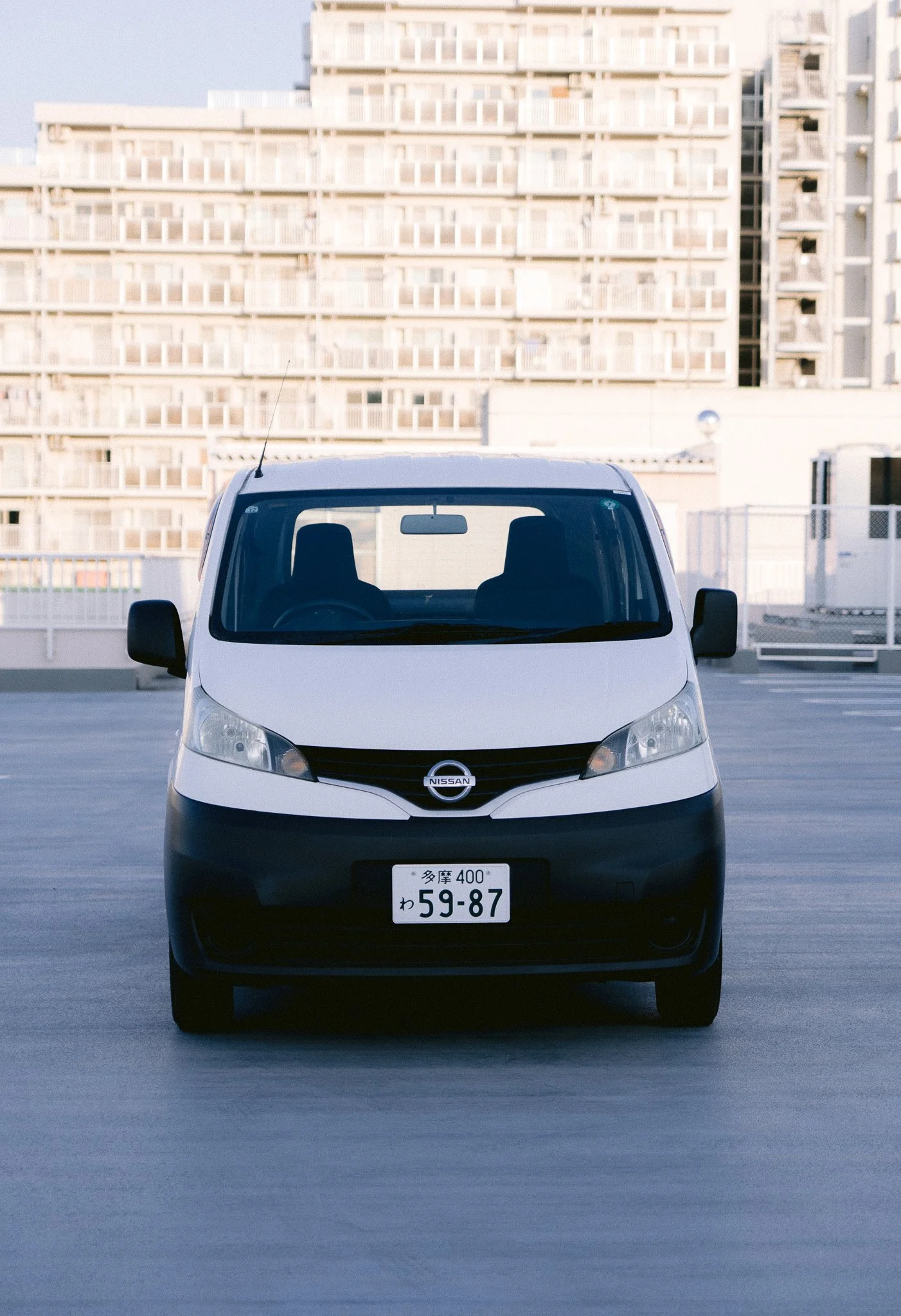 白いニッサンの軽自動車が駐車された高層マンションの屋上の駐車場。背景に多くのベランダのあるマンションの建物が見える。