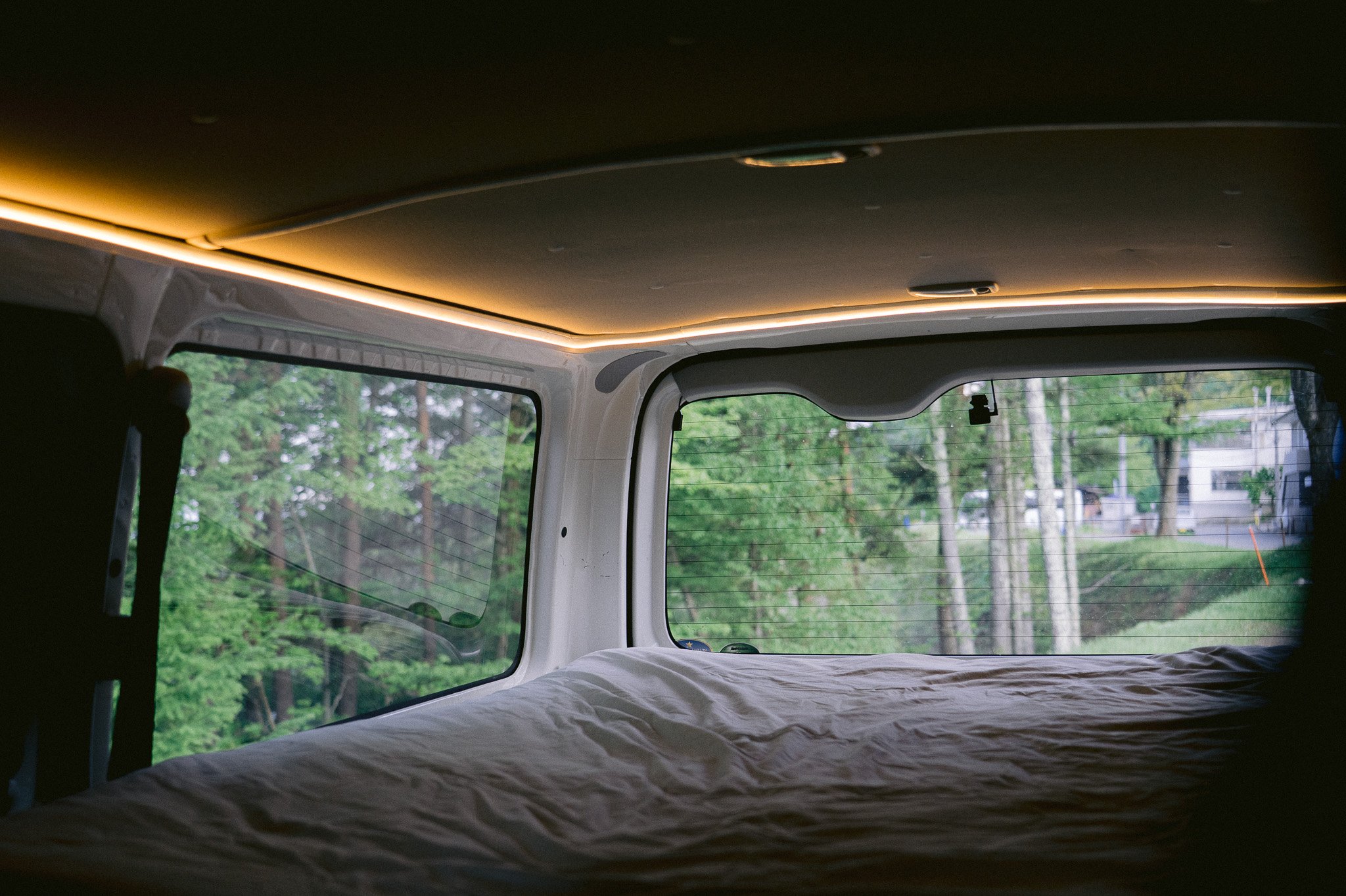 Campervan interior at night — a made bed looking out through the rear window onto a green forest