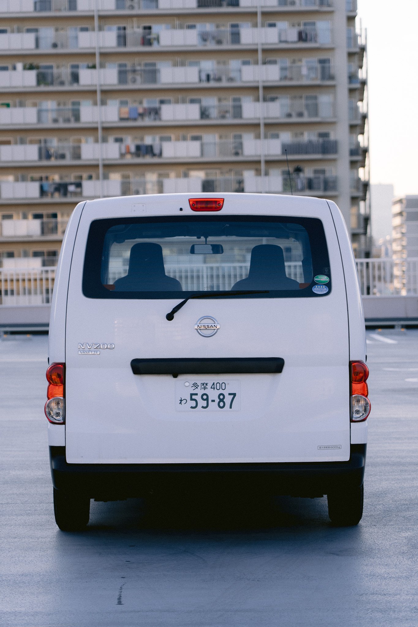 白いニッサンのバネットバンのリアビューと多摩市のナンバープレート、背景は高層アパートの建物