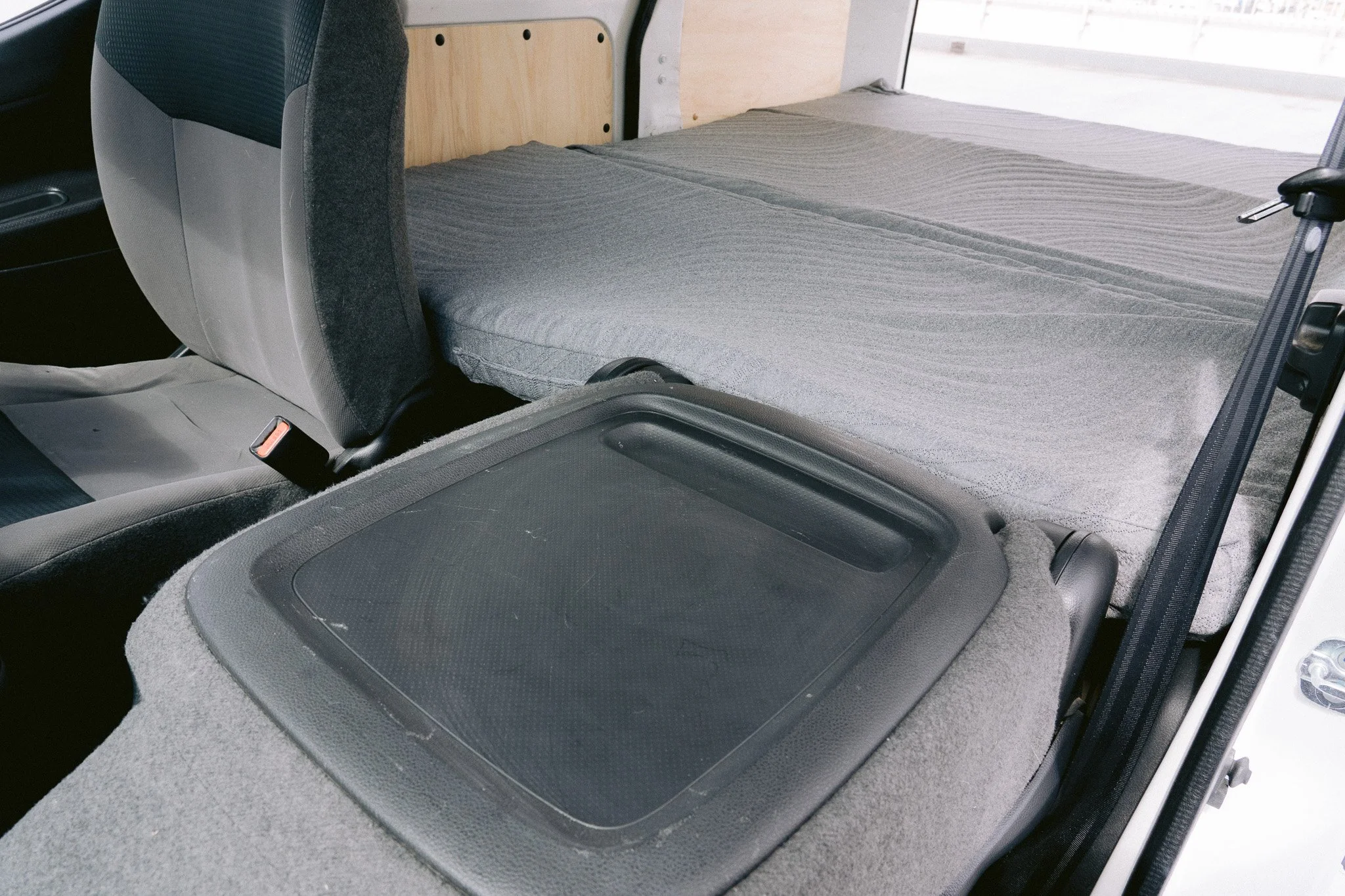 車の後部座席のフラットなベッドスペースに蓋付きの収納トレイと灰色のシートが見える車内の写真