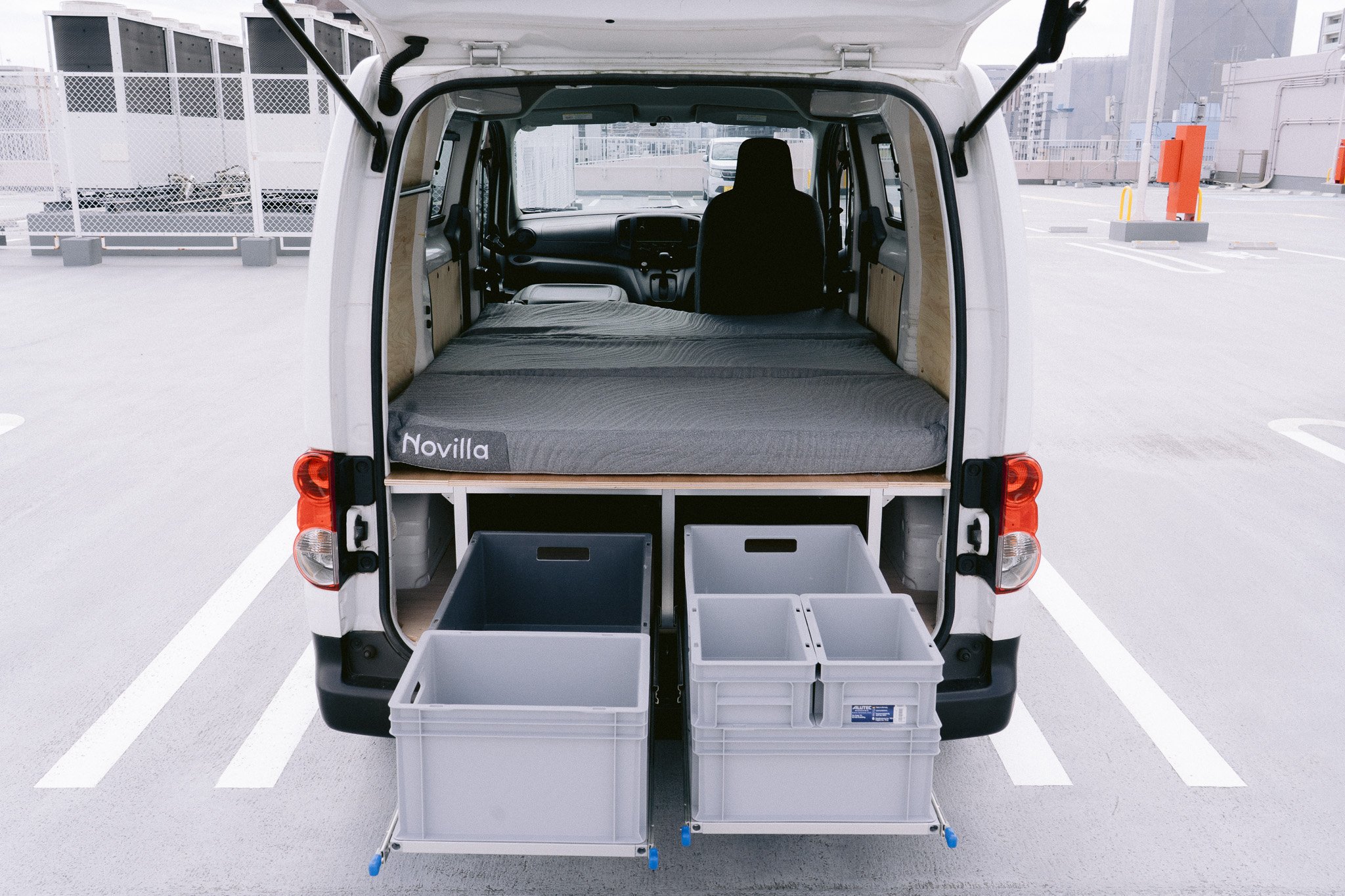 白いバンの後部が開かれ、荷物を置くための収納スペースといくつかのプラスチックケースが見える。車は屋上駐車場に停車している。