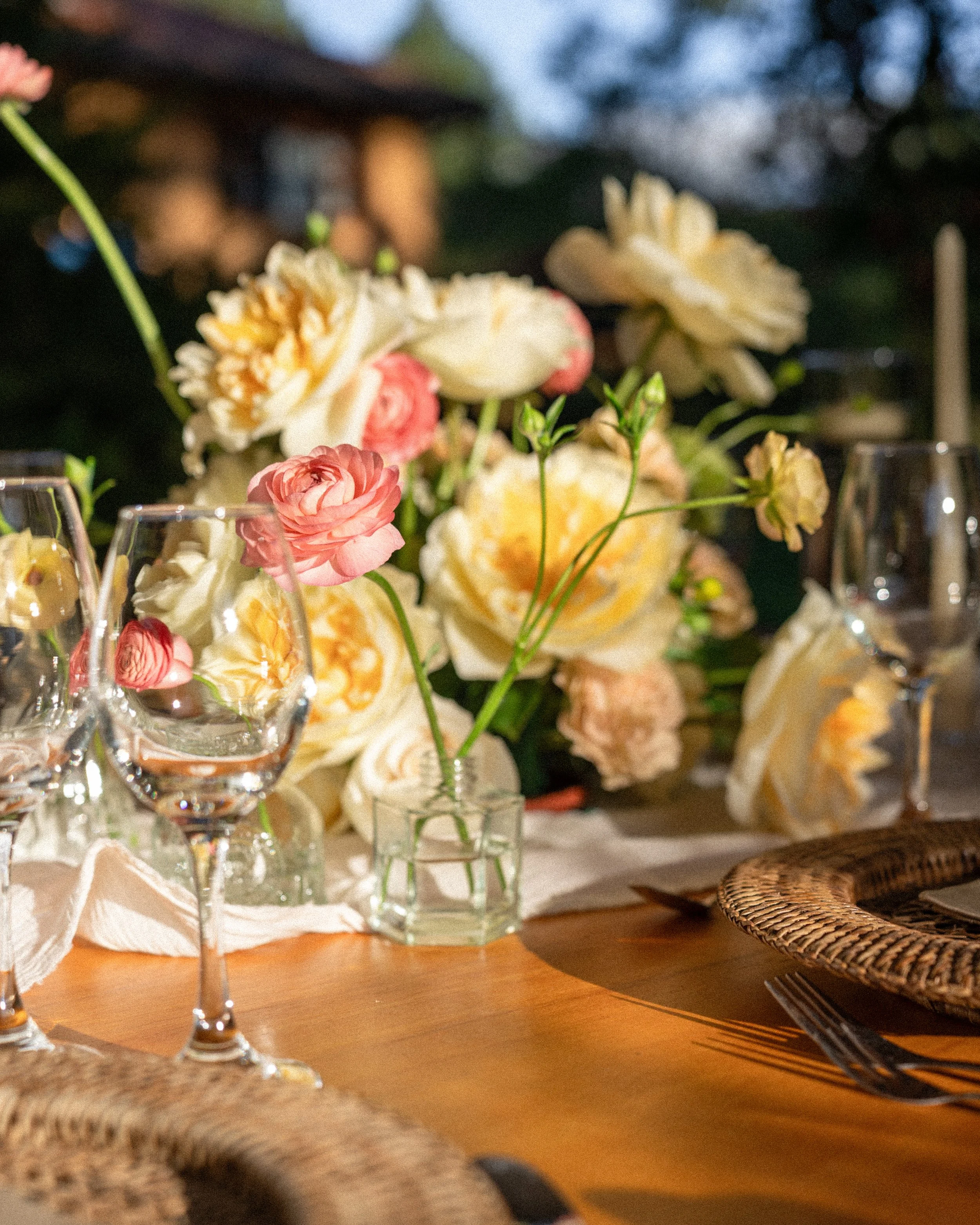 Boda Ale y Robby, Bautizo Carlota, arreglos florales por DAGA Studio. Mesa decorada con un arreglo floral de flores rosadas y blancas, varias copas de vino y cubiertos de madera en un ambiente al aire libre por la tarde.