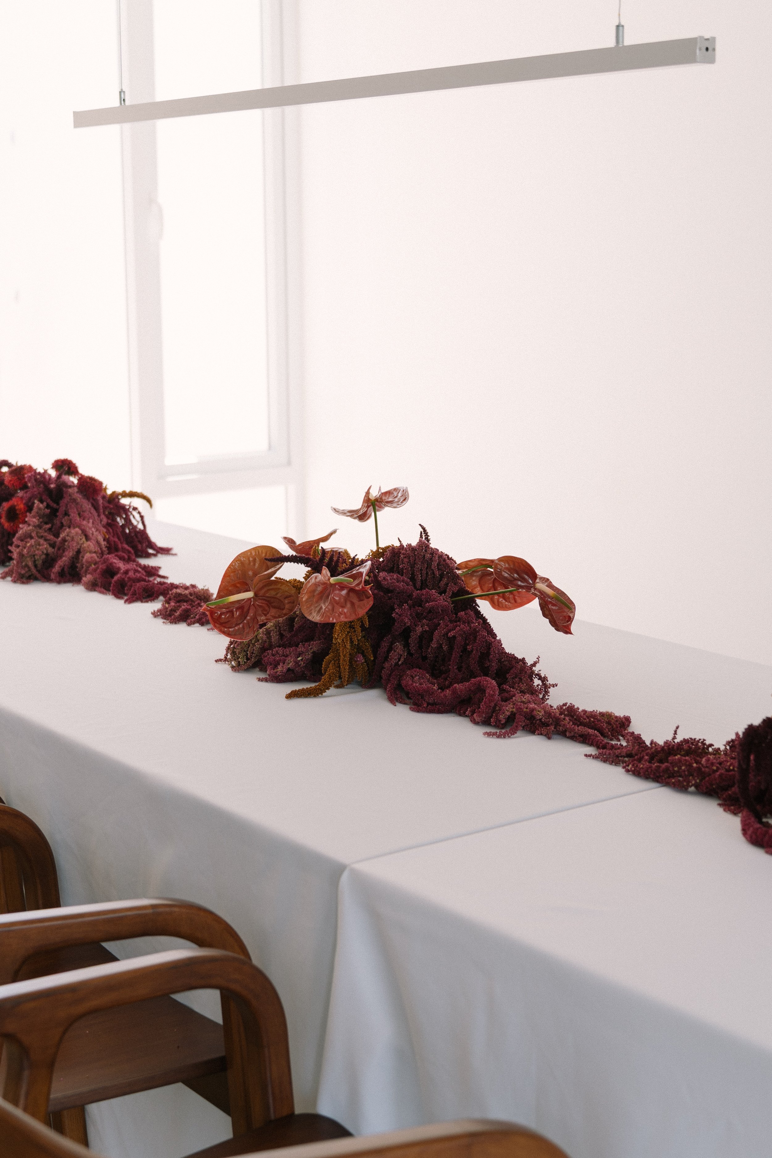 Boda Carolina y Juanjo, arreglos florales por DAGA Studio. Decoración con flores y hojas en una mesa blanca, en un espacio con paredes blancas y sillas de madera.
