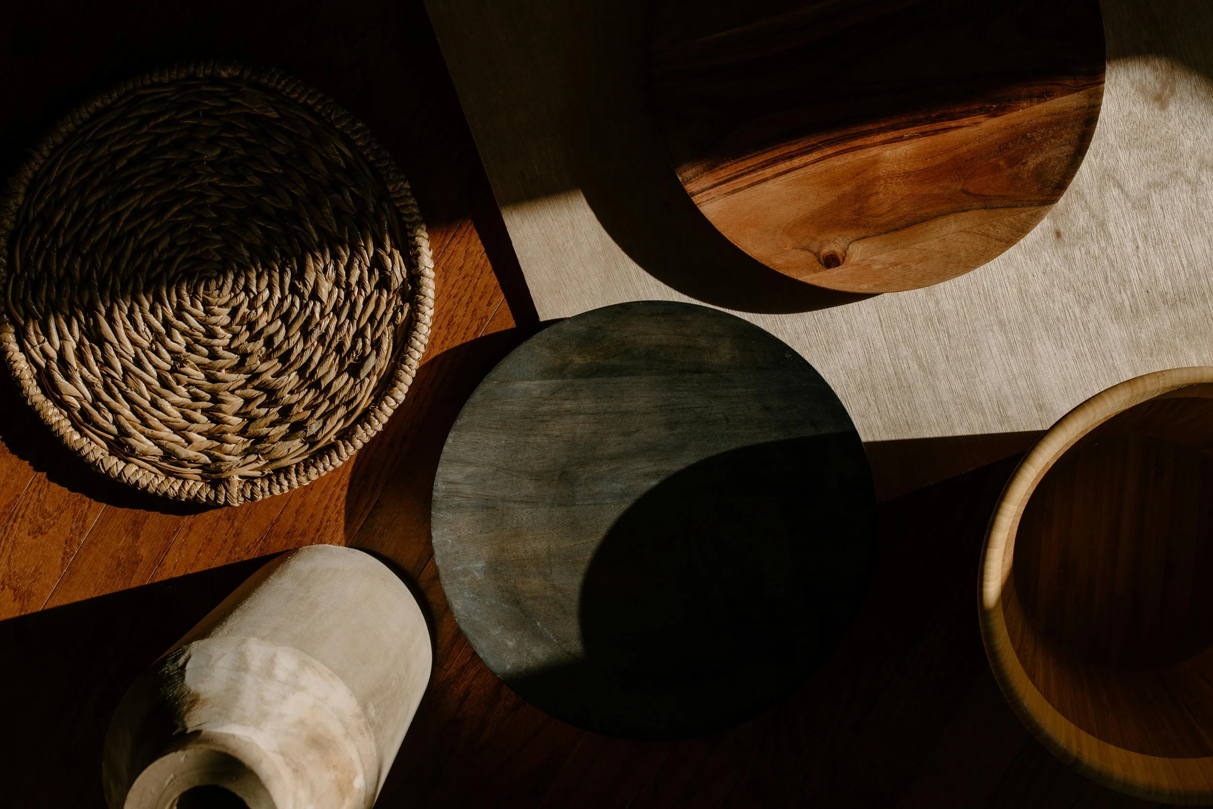 Top-down view of various wooden and woven decorative objects, including baskets and bowls, with sunlight casting shadows on a wooden surface.