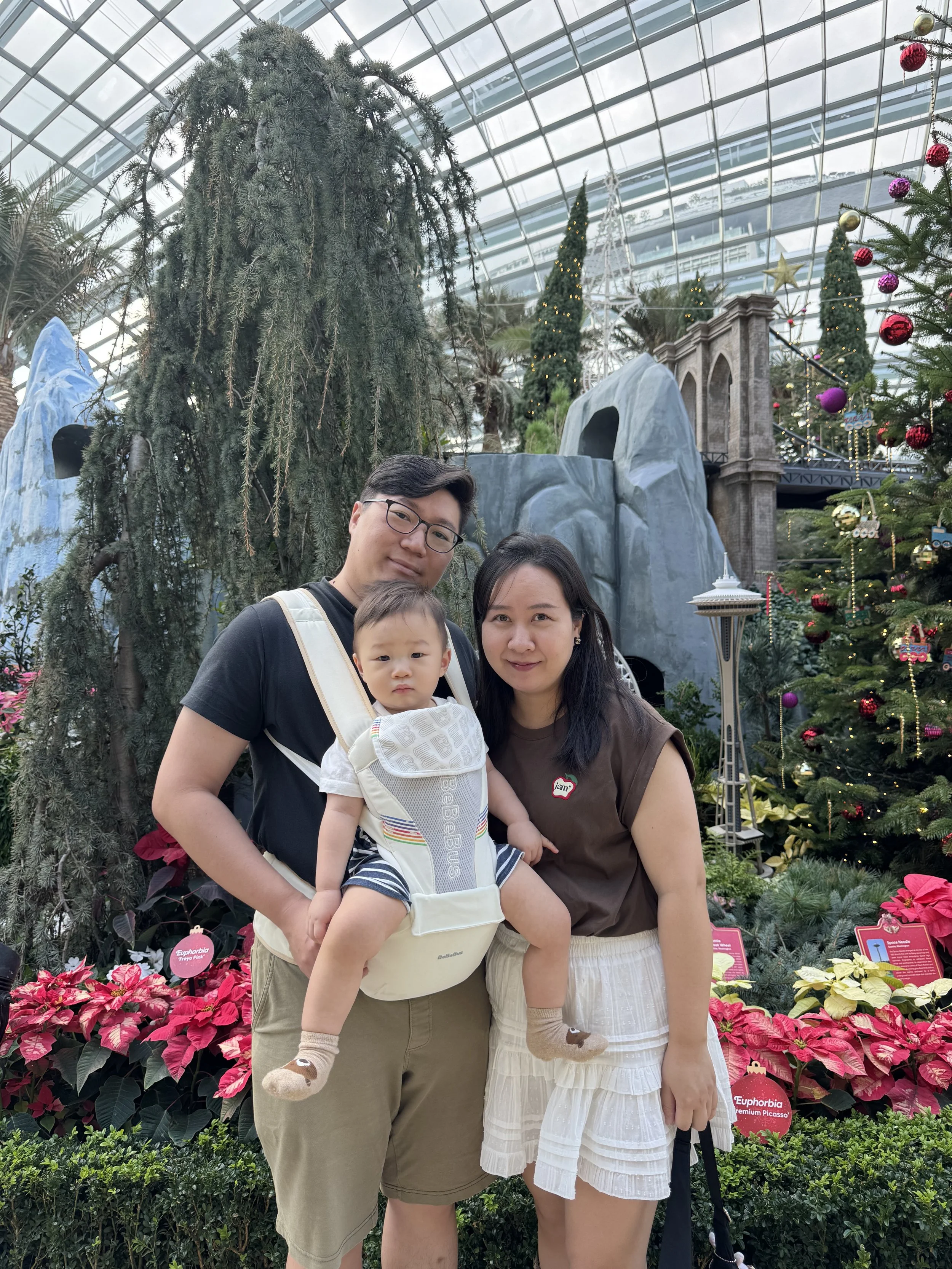 A family of three, including a man, woman, and young boy, standing in front of Christmas trees with ornaments and poinsettias inside a glass-domed botanical garden.