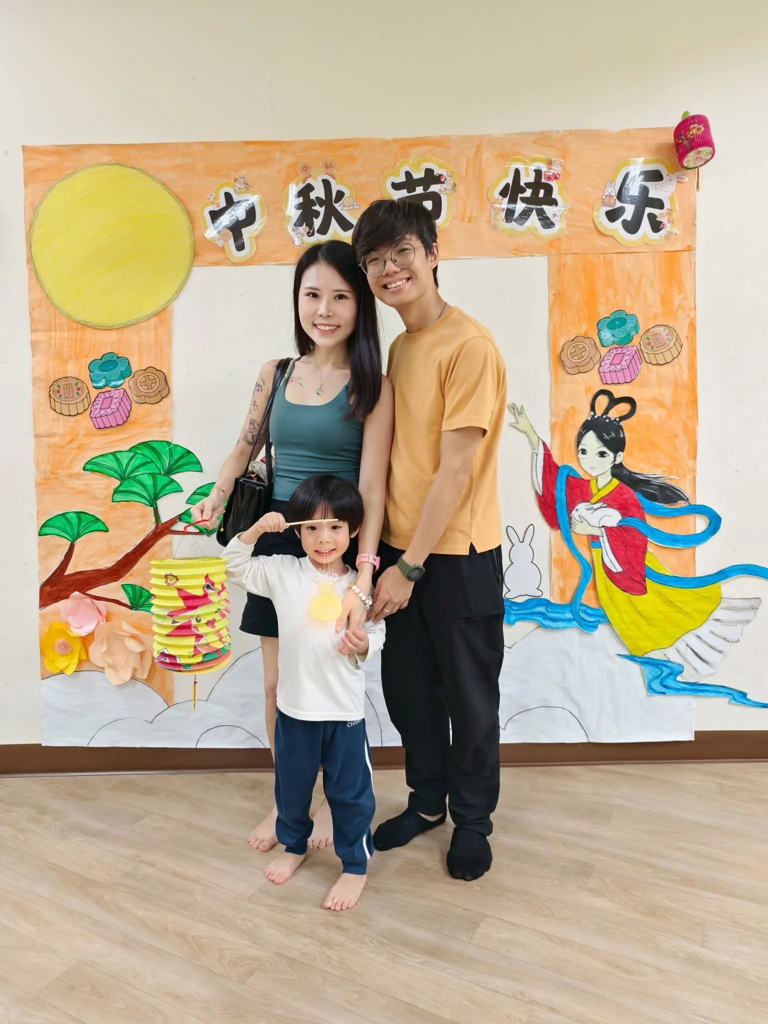 Family of three standing in front of a colorful backdrop with Chinese characters and moon-themed decorations for the Mid-Autumn Festival, including a large yellow moon, paper lanterns, and a traditional lady figure.