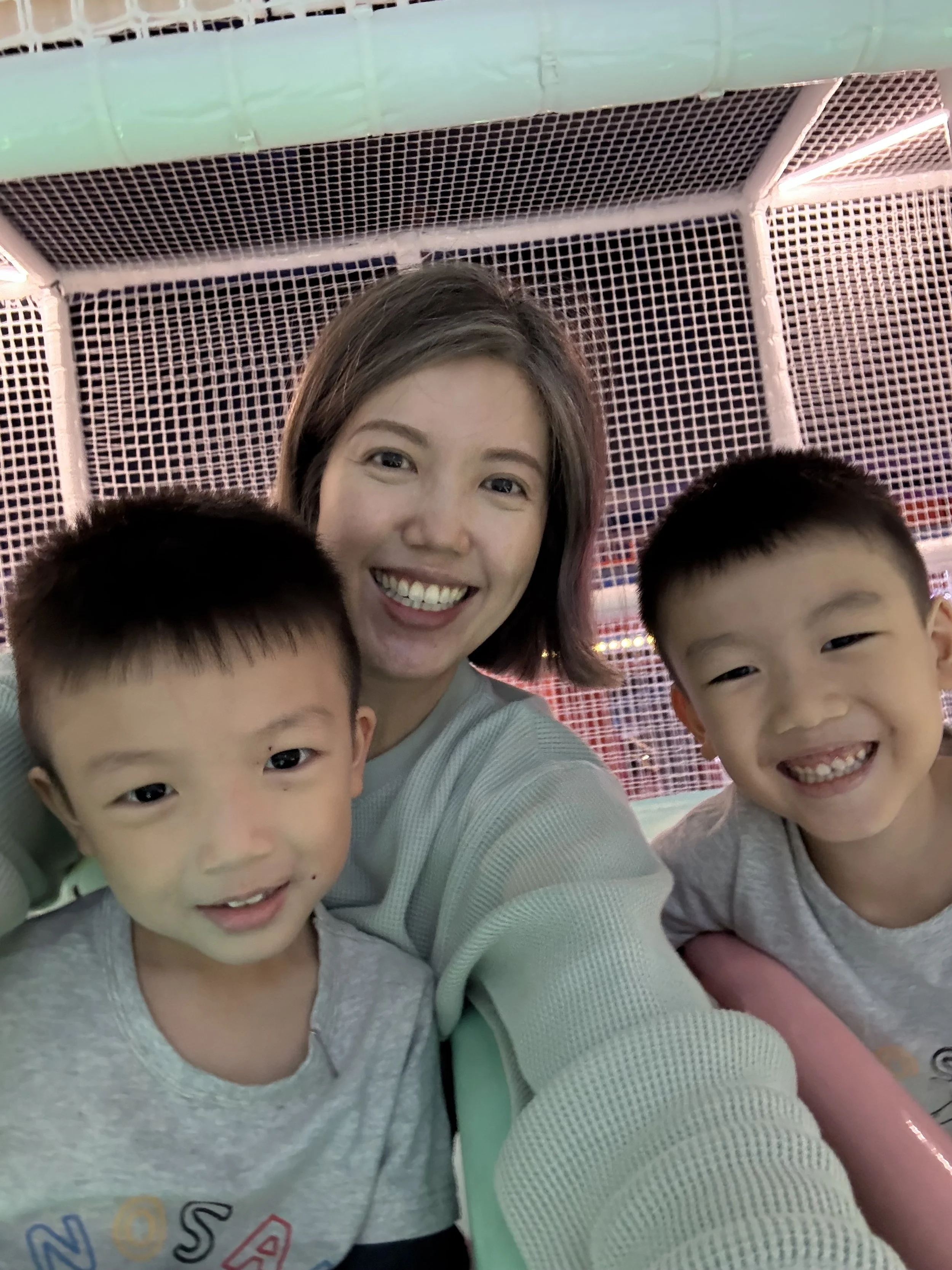 A woman and two young boys taking a selfie together inside a brightly colored amusement park ride with a pink and green netted background.