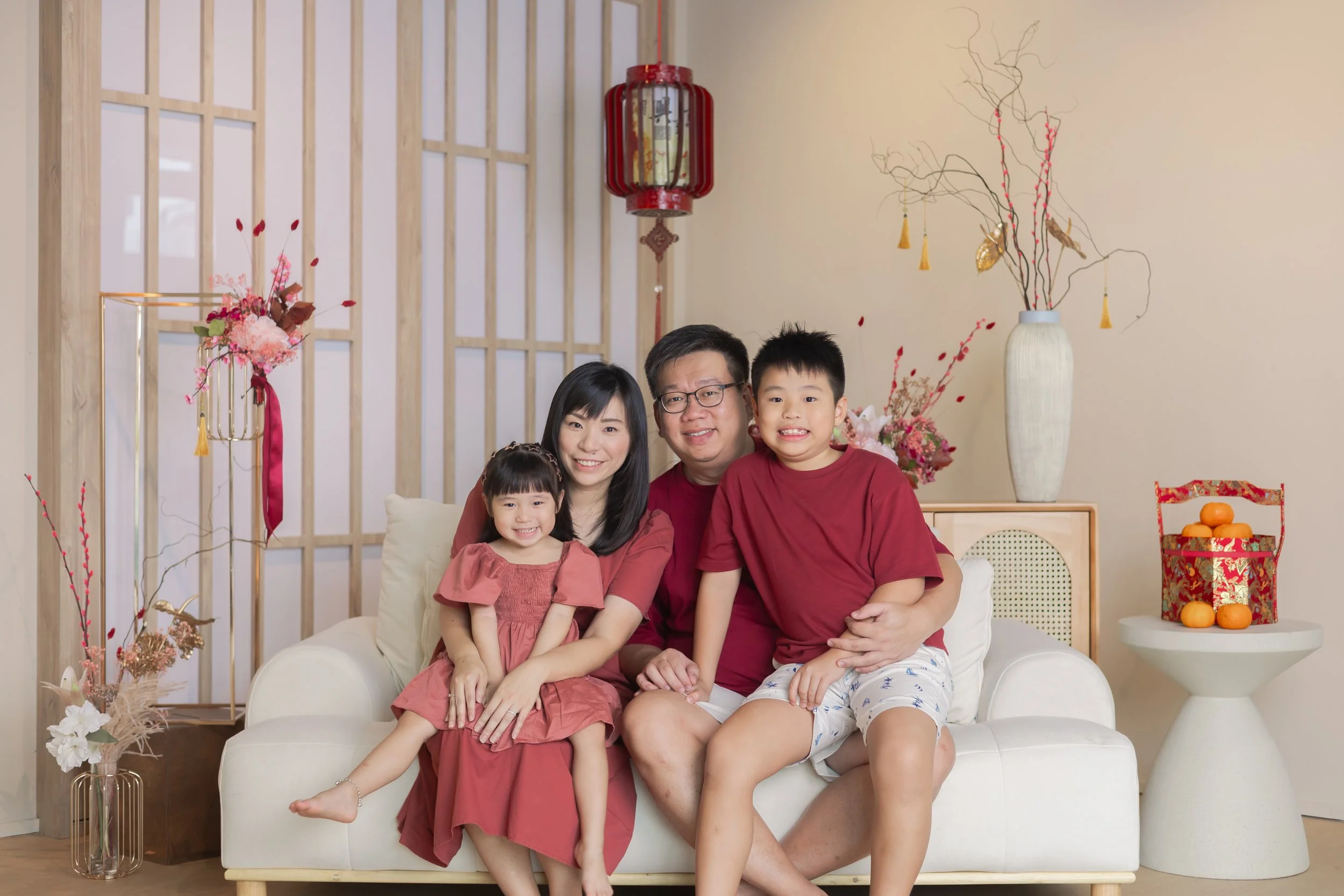 Family of four sitting on a white couch with Asian features, celebrating Lunar New Year. The family includes a father, mother, son, and daughter, all wearing red. The room is decorated with traditional Lunar New Year decorations, including large vases with floral arrangements, a red lantern, and a small table with oranges. The background features wooden lattice panels and beige walls.