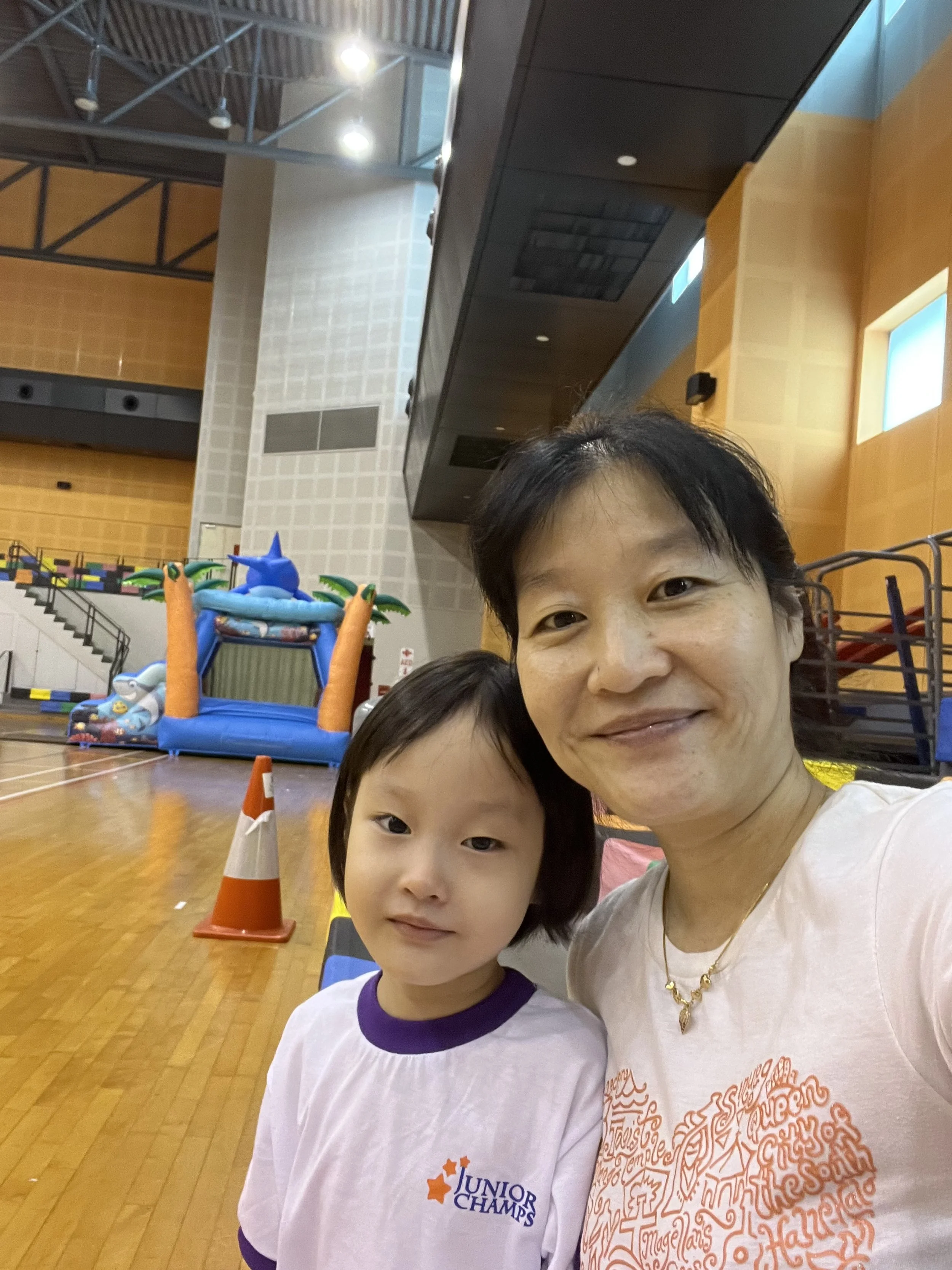 A woman and a young girl taking a selfie inside a gymnasium or indoor sports facility. In the background, there is a colorful inflatable bounce house and some traffic cones on the wooden floor.