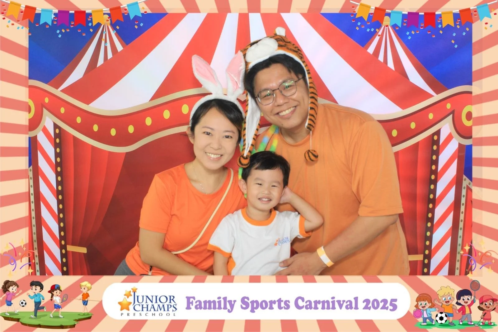 A family of three, wearing orange shirts, poses for a photo at the Junior Champs Family Sports Carnival 2025. The mother and father are both wearing animal-themed headgear, with the mother in bunny ears and the father in a tiger hat. They are smiling and standing in front of a colorful circus-themed backdrop featuring red and white stripes, flags, and confetti. The young boy in front is smiling, wearing a white shirt with orange accents, and has his hand behind his head.