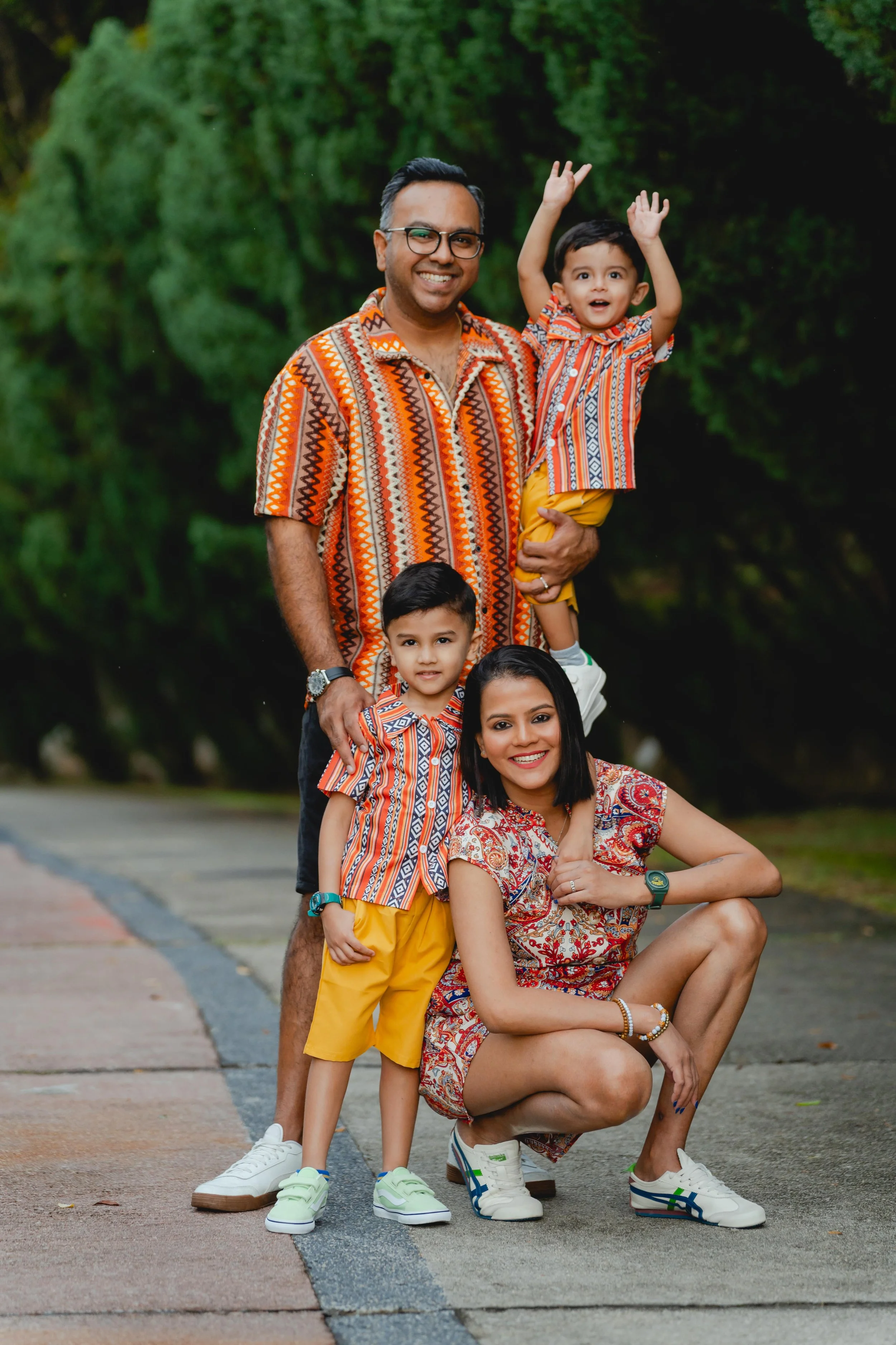 Family of four posing outdoors, middle-aged man and woman with two young boys, all wearing colorful patterned attire, standing on a paved path with green foliage in the background.