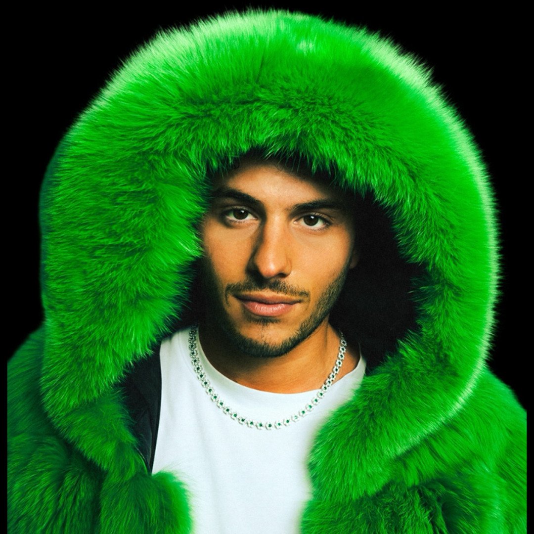Portrait of a young man wearing a large, bright green fur hooded coat against a black background.