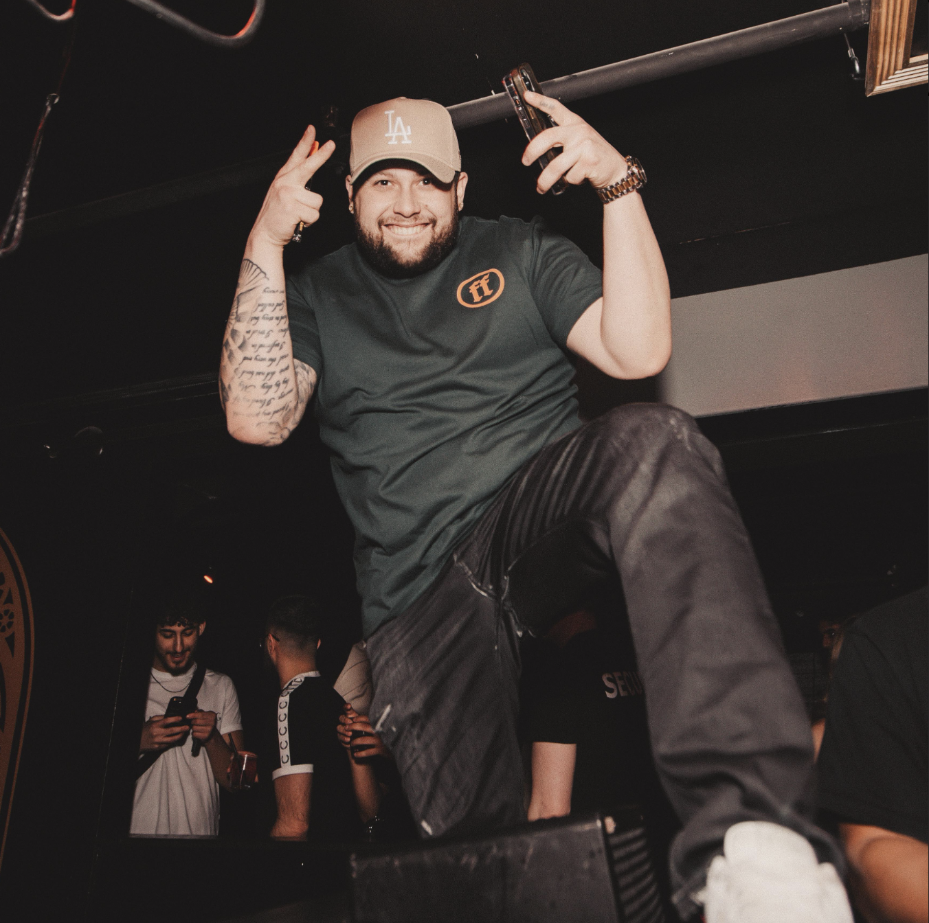 A man with a beard, wearing a beige cap, black t-shirt, and jeans, smiling, making peace signs, holding a phone and a drink, standing on a raised surface in a club or party setting with other people in the background.