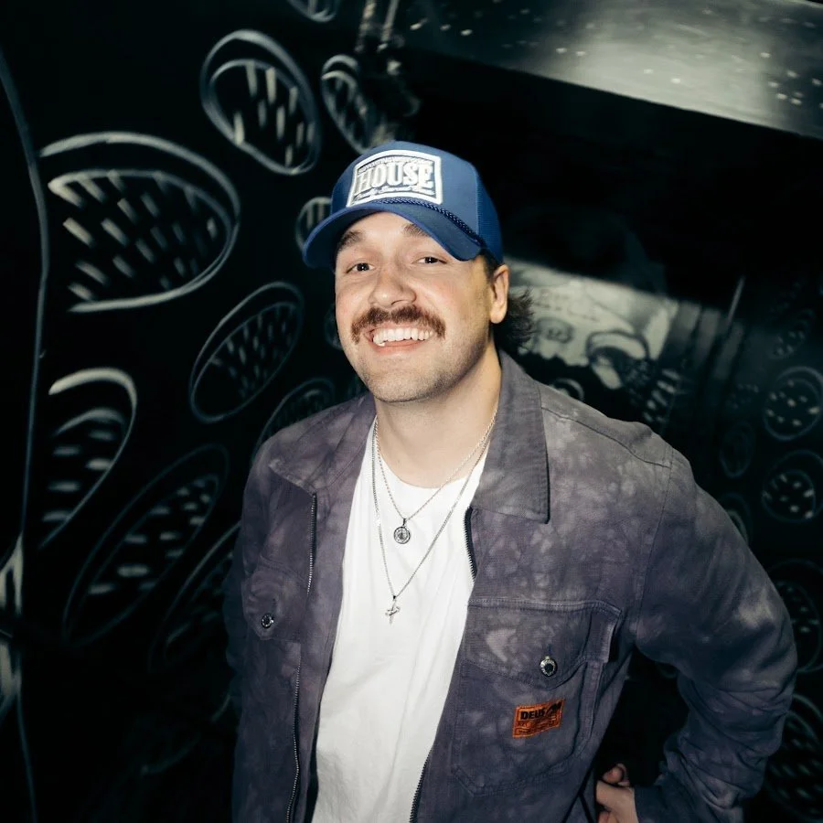 A smiling man with a mustache and beard wearing a blue cap, gray jacket, and white shirt, standing in front of a black wall with white oval patterns.