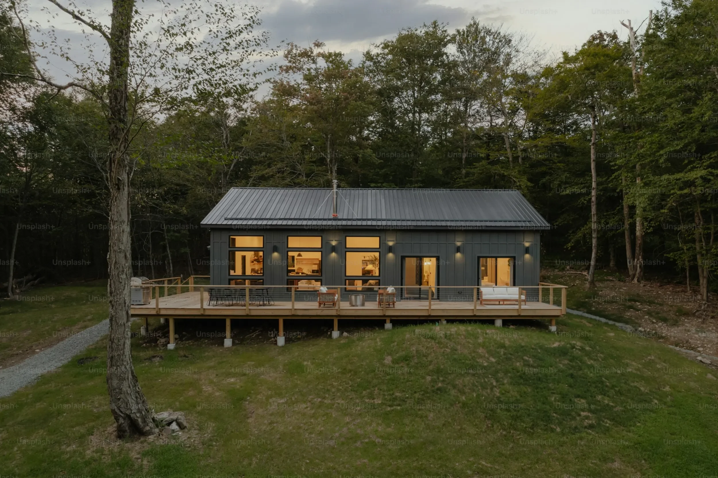 A modern house with large windows and a metal roof, situated on a grassy hillside surrounded by trees during dusk, with warm interior lighting visible through the windows.