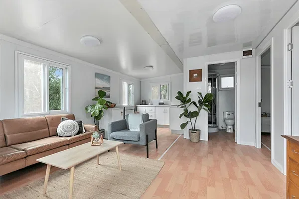 Living room with a brown sofa, gray armchair, coffee table, and potted plants, with windows showing greenery outside and a small kitchen area in the background.