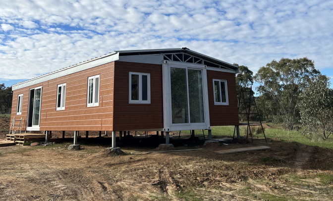 A tiny house under construction on stilts with brown siding and large windows, set in a rural area with trees and a partly cloudy sky.