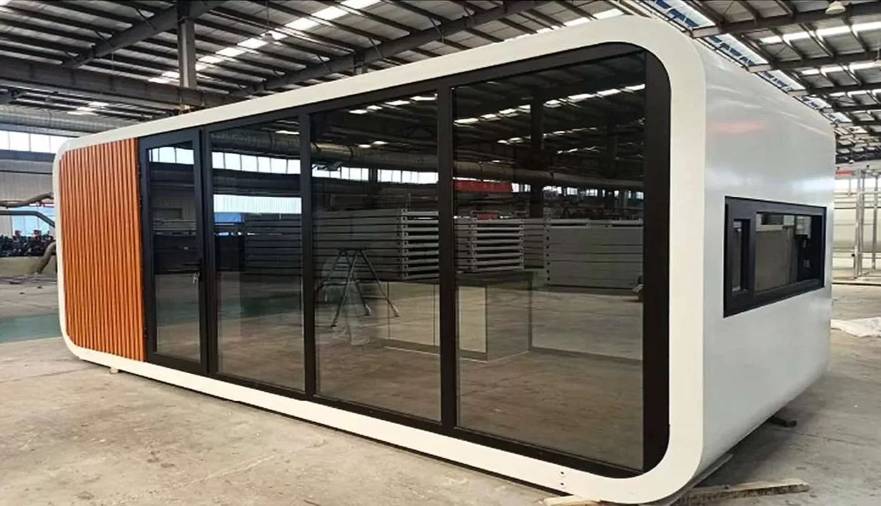 A modern, oval-shaped prefabricated building with large glass sliding doors and a section of vertical wooden slats on one side, inside a warehouse.