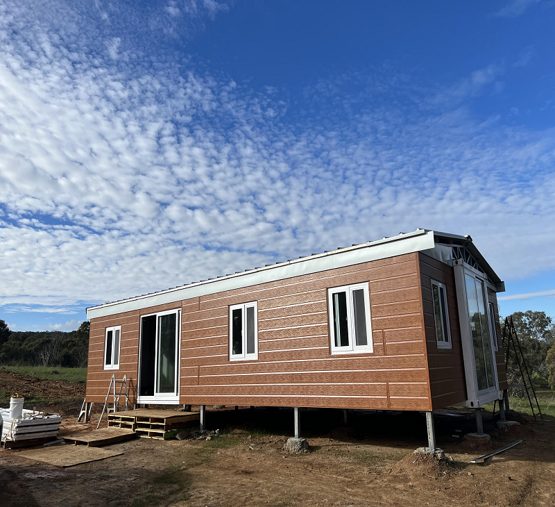 A tiny, pre-fabricated house with wood exterior siding, multiple windows, and a small set of stairs leading to a door, set outdoors on stilts on a clear day with scattered clouds.