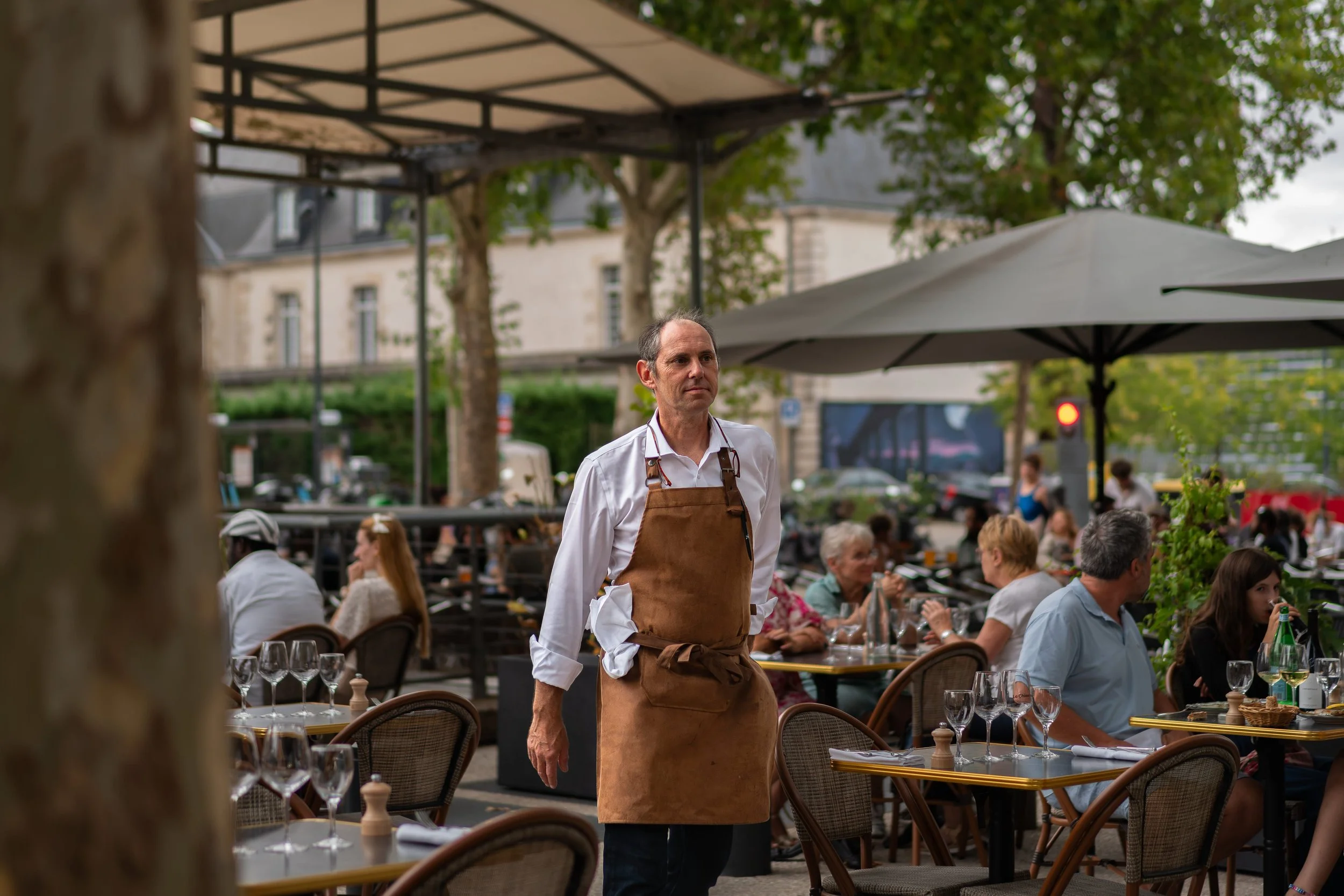 Un homme debout, portant un tablier en cuir et une chemise blanche, dans un restaurant en plein air remplie de clients. Arbres et bâtiments en arrière-plan.
