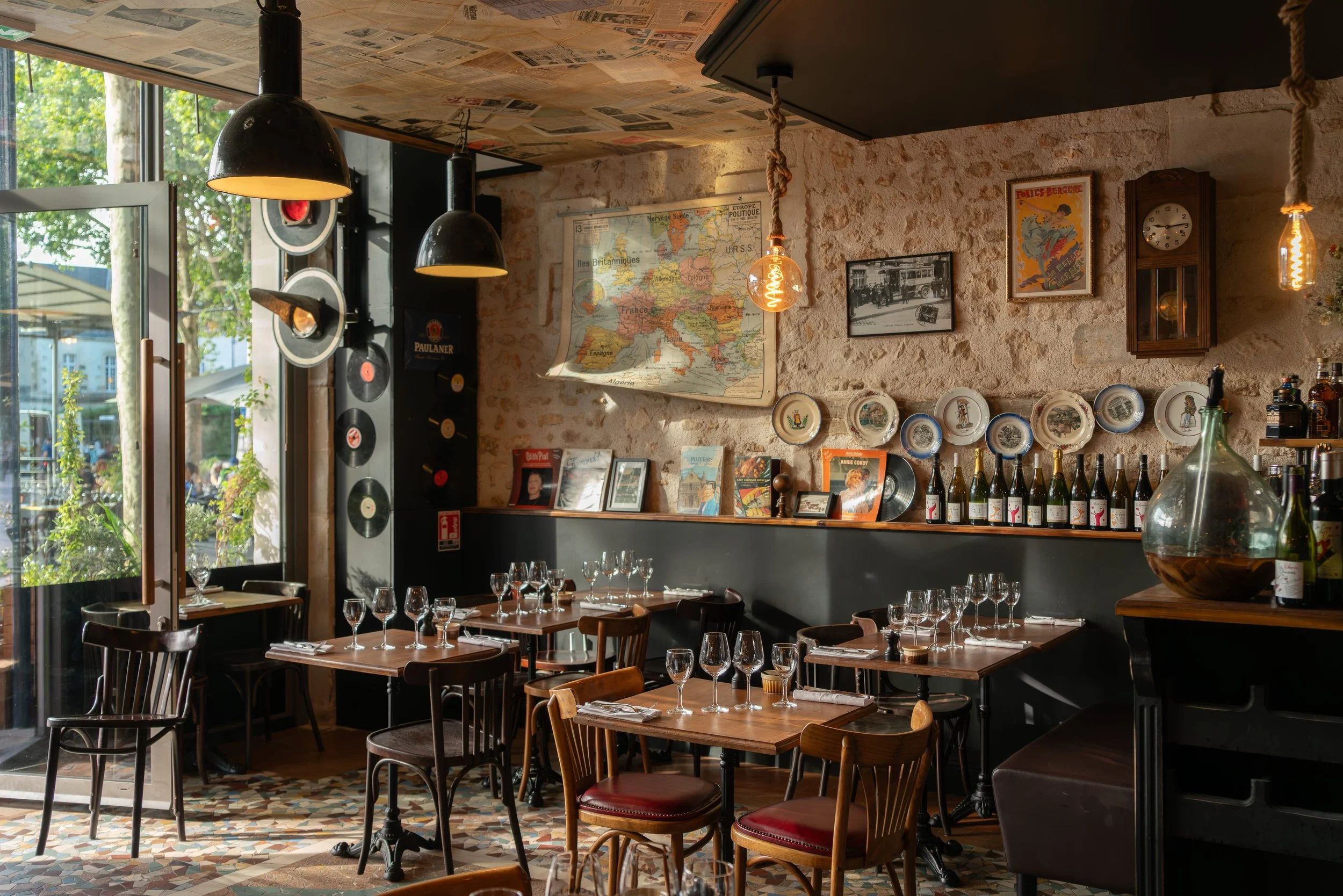 Intérieur d'un restaurant avec des tables en bois et des assiettes en porcelaine, bouteilles de vin alignées sur un meuble en arrière-plan, murs en pierre avec décor vintage, lampes suspendues avec ampoules Edison, une grande fenêtre laissant entrer 