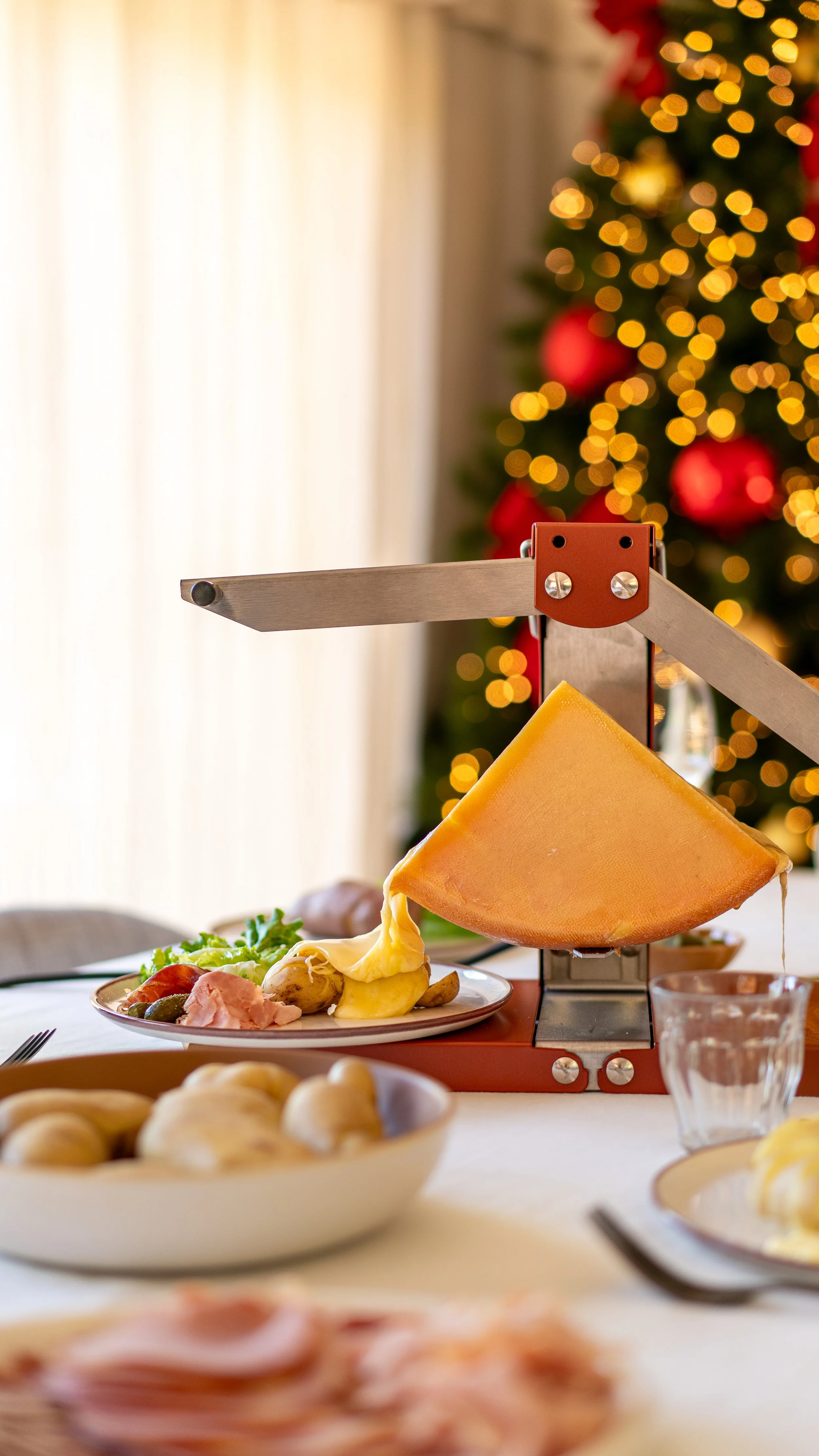 Un plateau avec du fromage, des légumes, du jambon, et autres aliments, avec un arbre de Noël décoré en arrière-plan, illuminé de lumières jaunes.