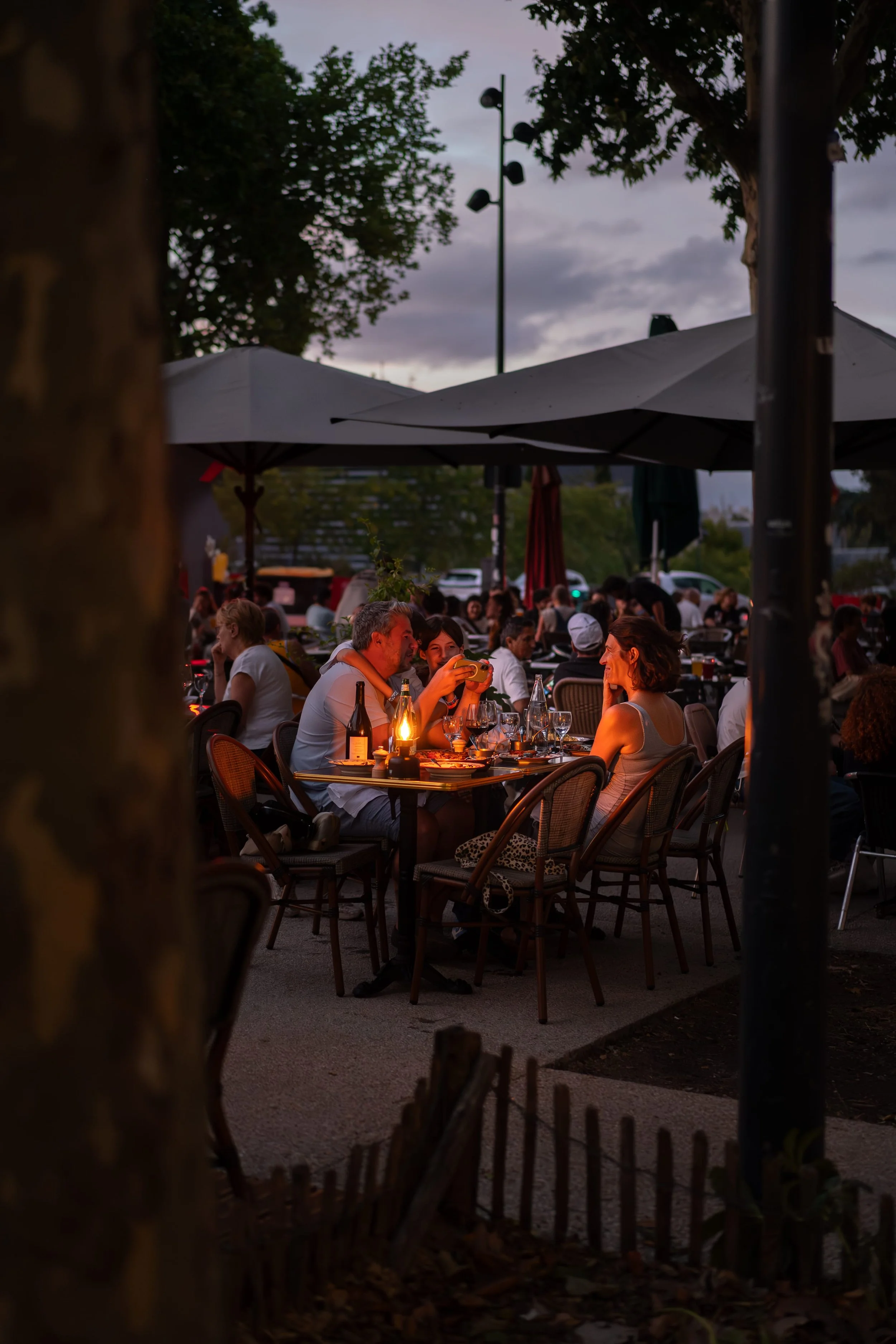 Un groupe de personnes assises à une terrasse de restaurant en soirée, avec des parasols, une lanterne et des arbres en arrière-plan.