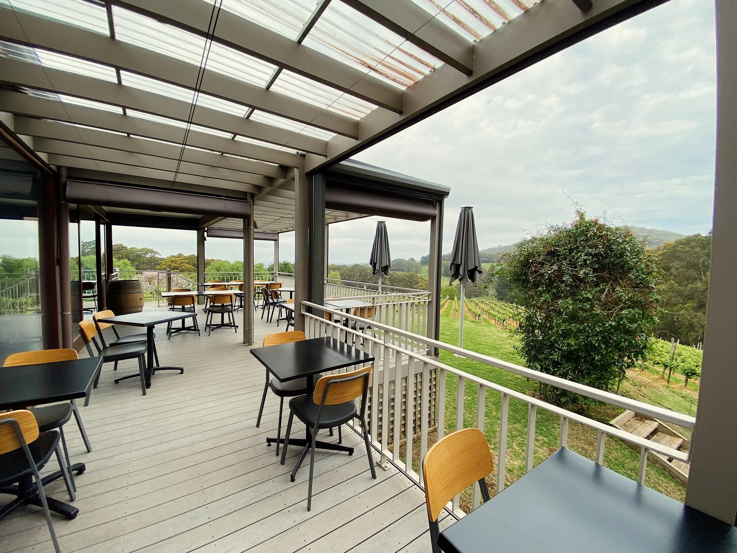 Empty outdoor patio with black tables and yellow chairs overlooking a vineyard and green landscape on a cloudy day.