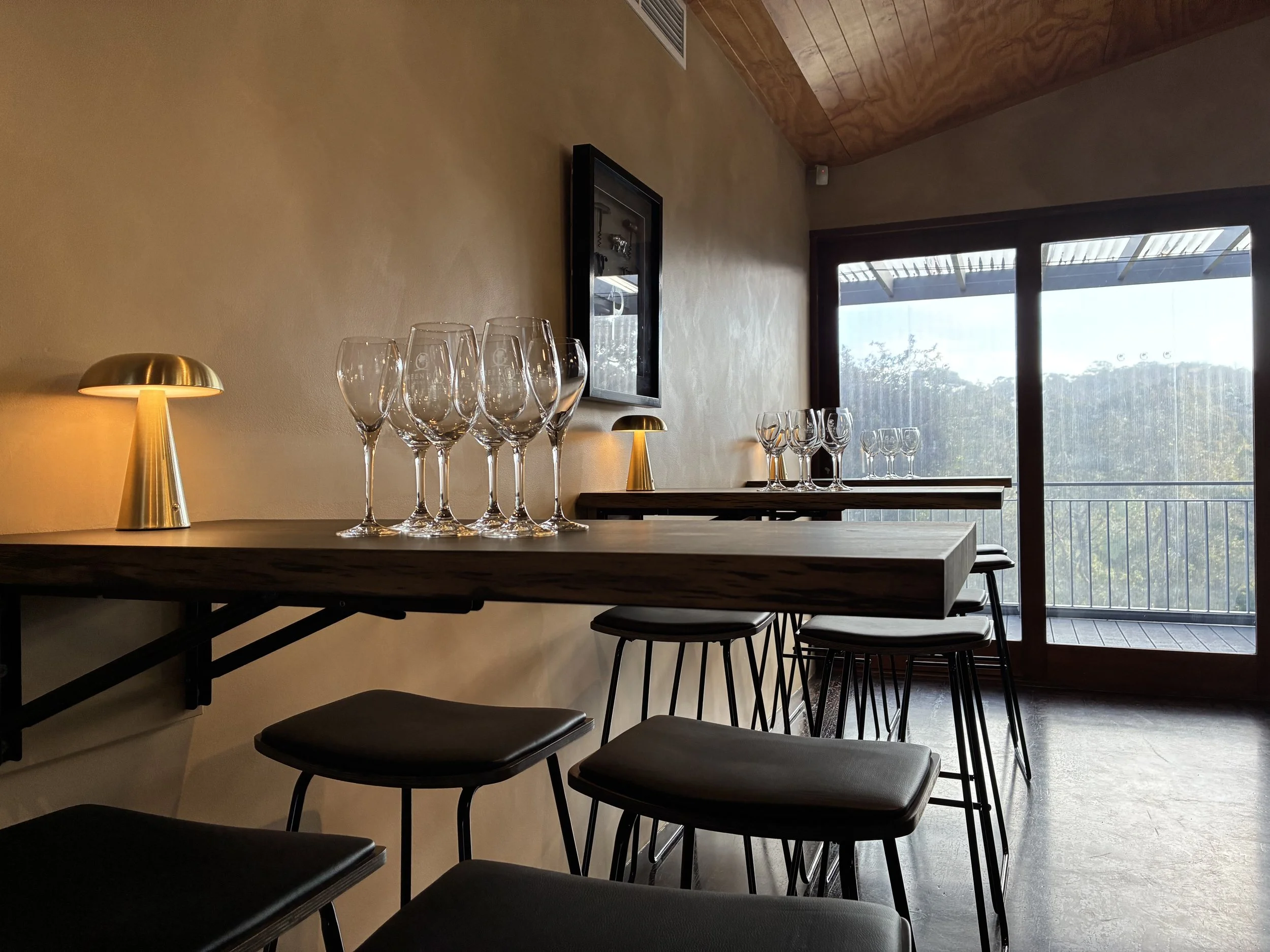 Interior of a dining area with a wooden counter and barstools, set with wine glasses and small lamps, overlooking a balcony through sliding glass doors with a view of trees and sky.