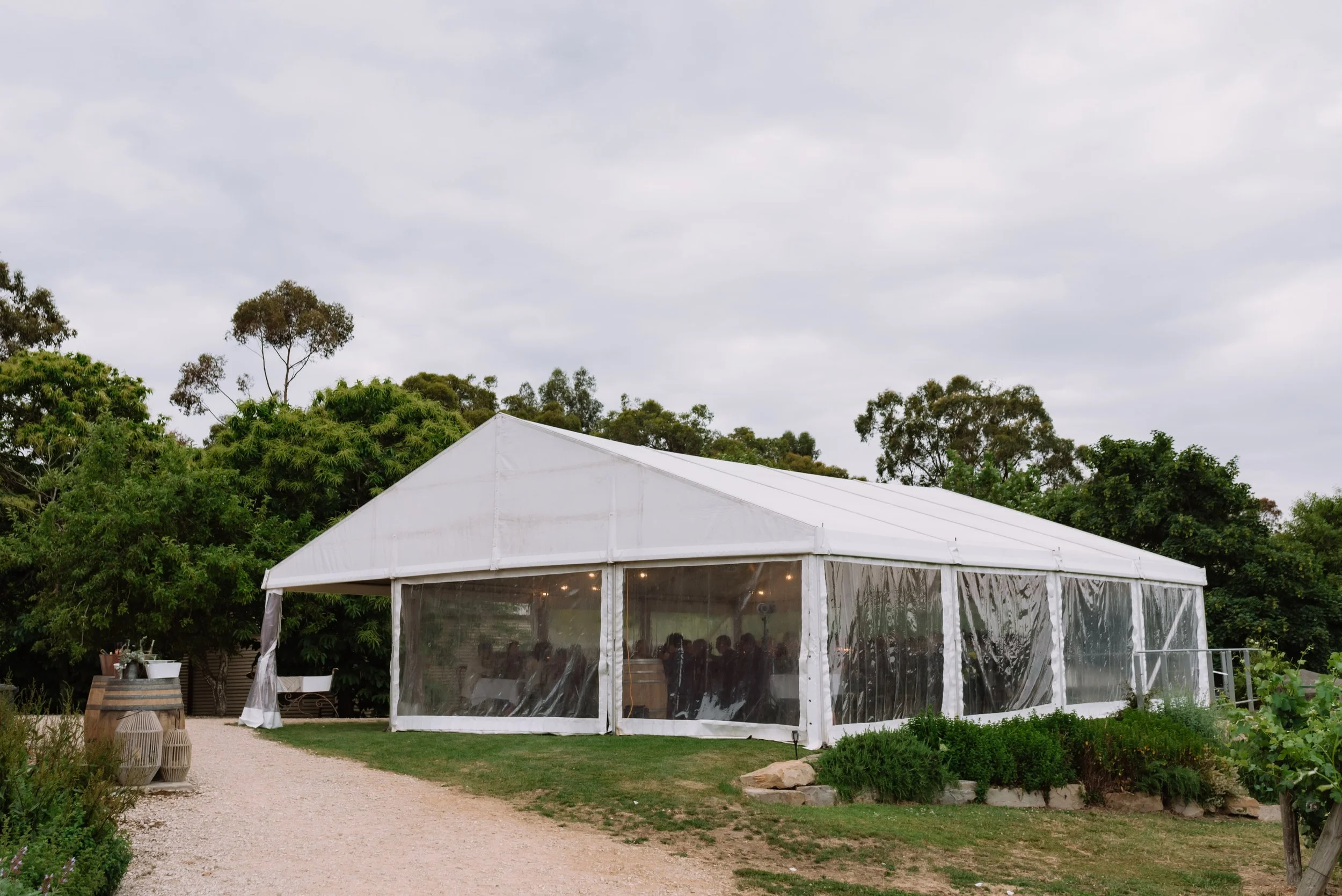 White event tent with transparent side walls on a grassy area, trees in the background, and cloudy sky overhead.