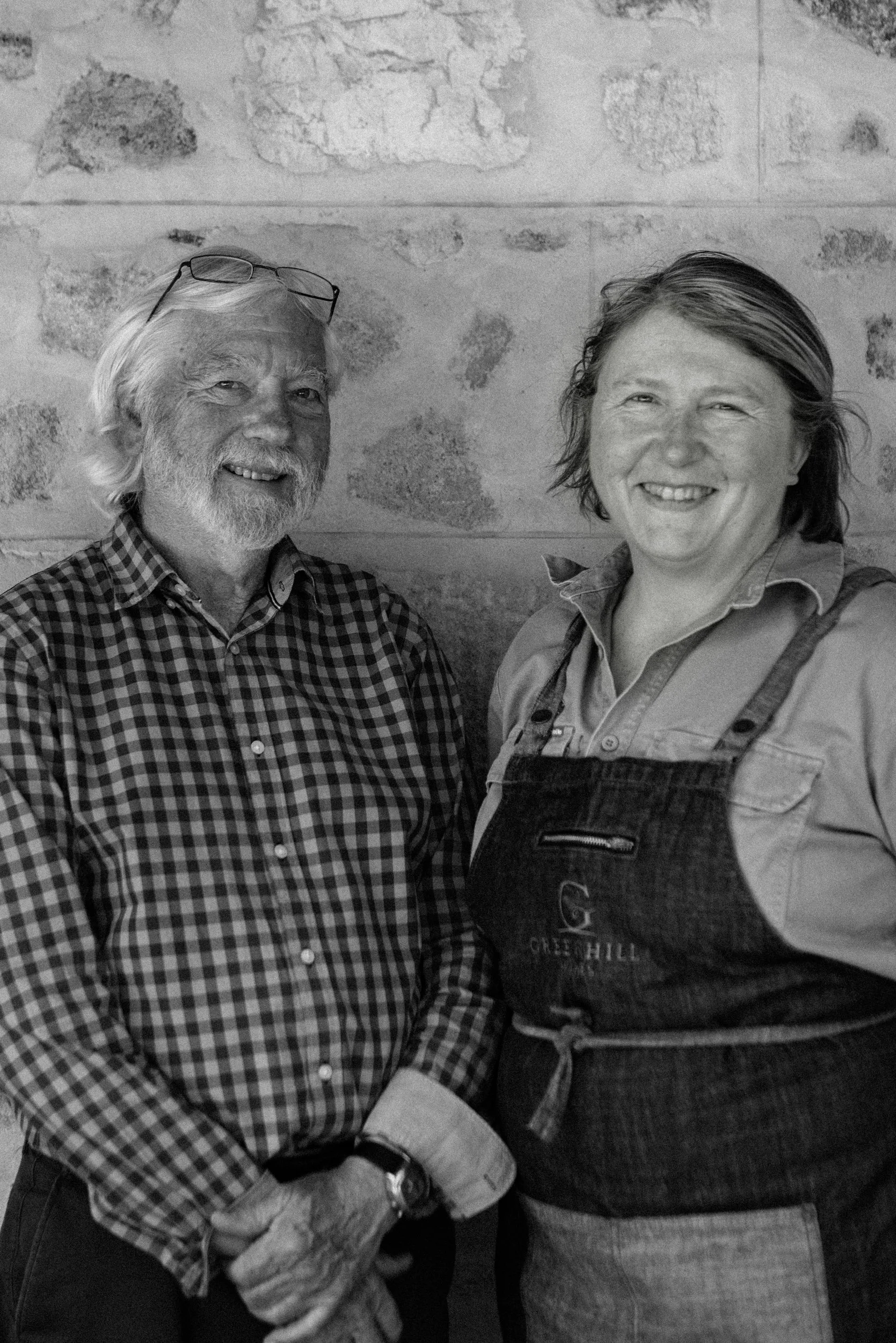 A black and white photo of an older man with glasses on his head, beard, and checkered shirt, standing next to a woman in a button-up shirt and apron, both smiling.
