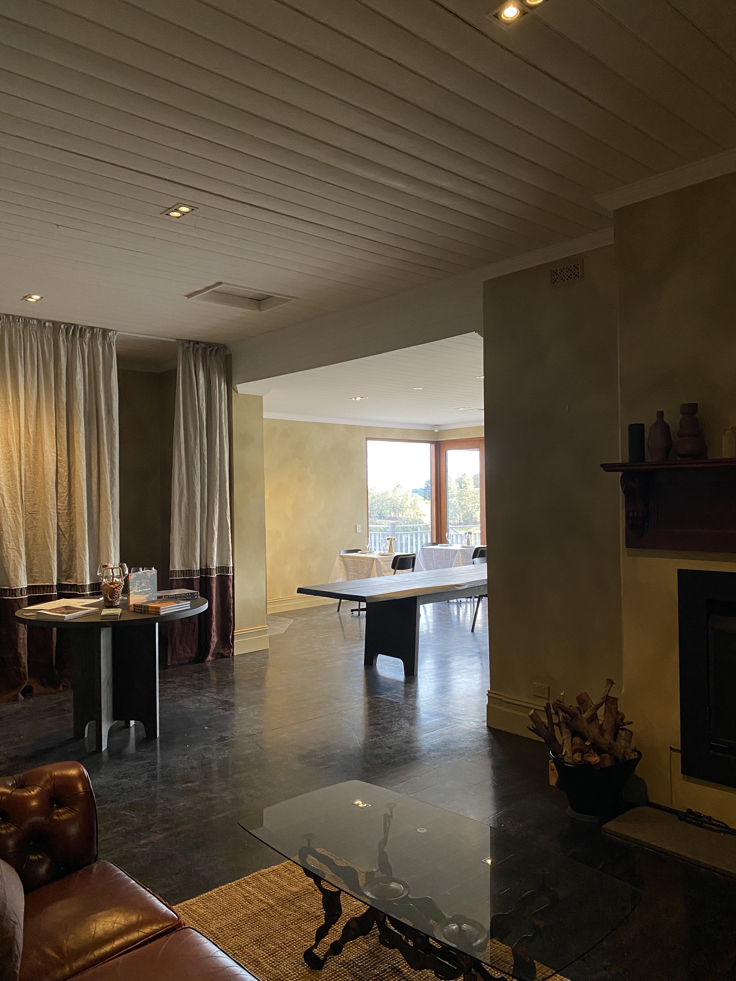 Living room with a leather sofa, glass coffee table, fireplace with a basket of firewood, and a section with dining tables near large windows.