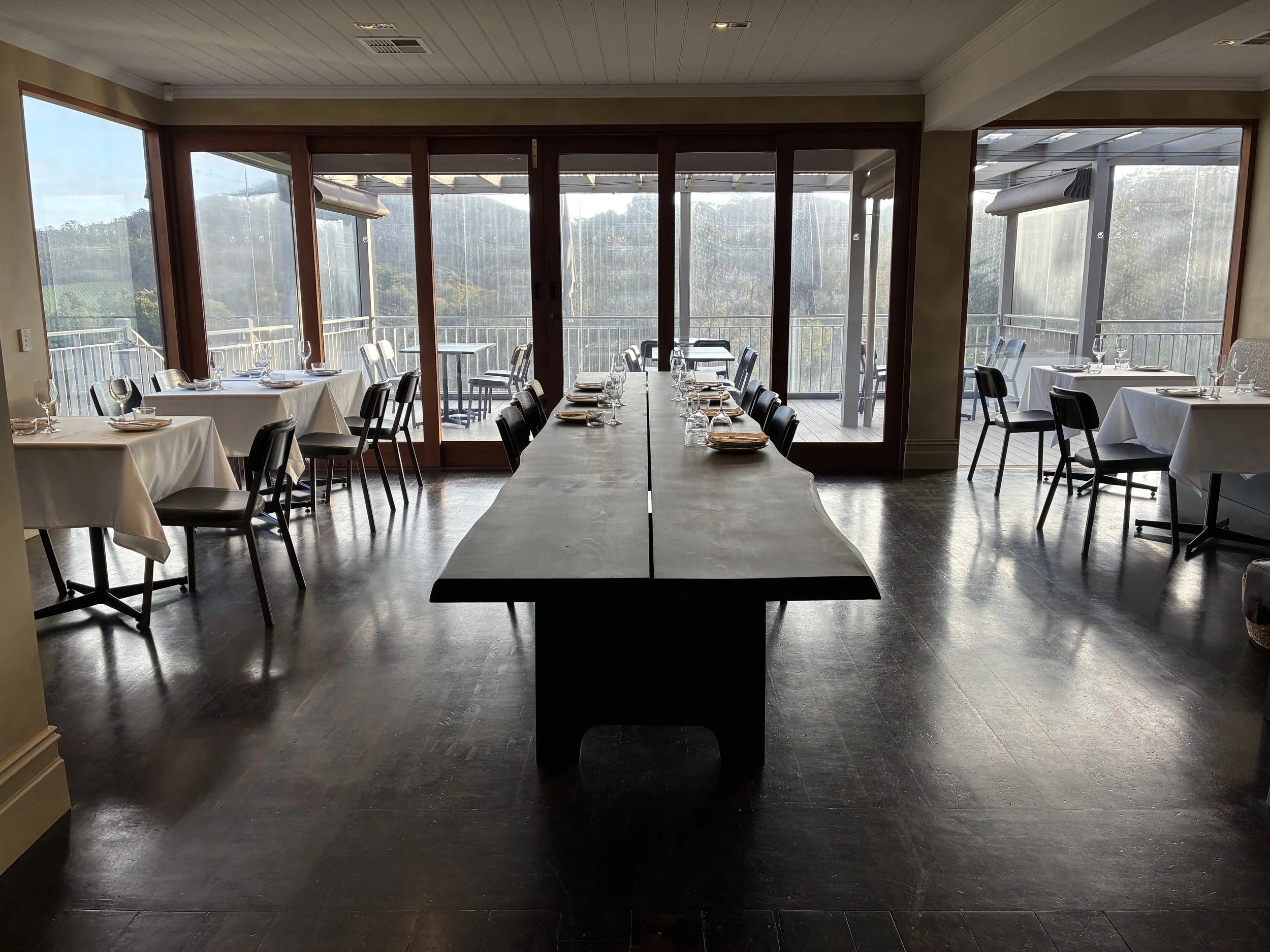 Restaurant dining area with white tablecloths, black chairs, and a large wooden table in the center, overlooking a balcony with outdoor seating and glass sliding doors.