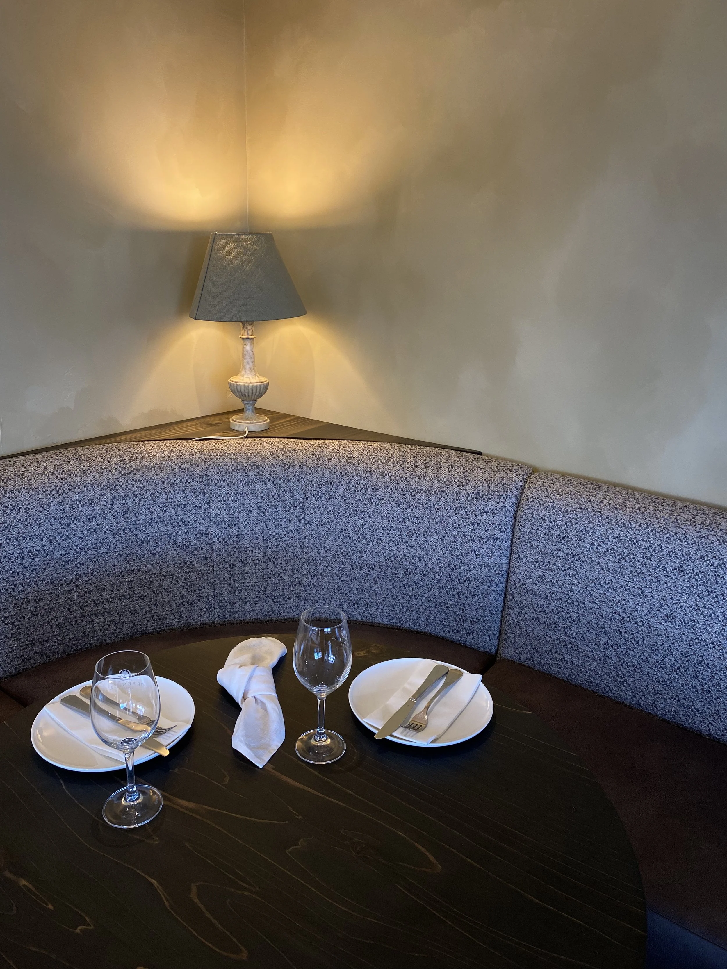 A round dining table set with two empty wine glasses, two white plates with cutlery, a folded napkin, and a beige cushioned banquette in a cozy corner, illuminated by a table lamp on the wall.