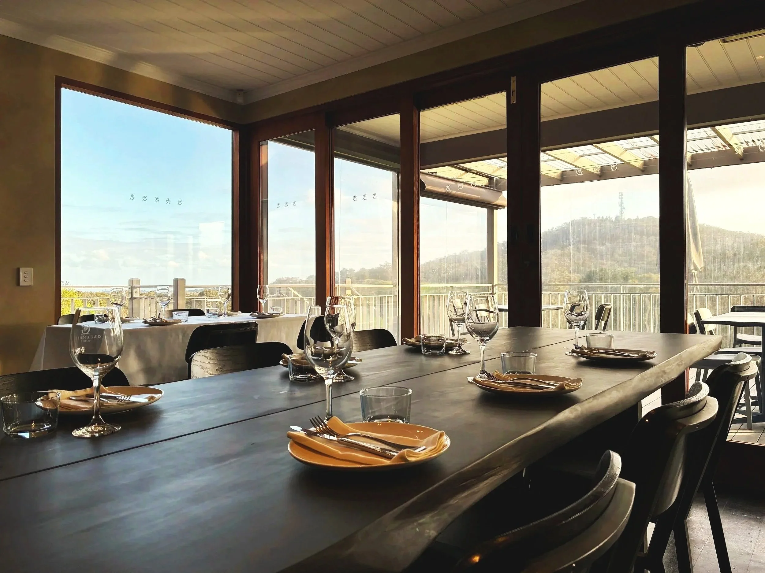 Indoor dining area with a long table set for a meal, featuring wine glasses, plates, and cutlery, overlooking a scenic outdoor view of mountains and sky through large windows.