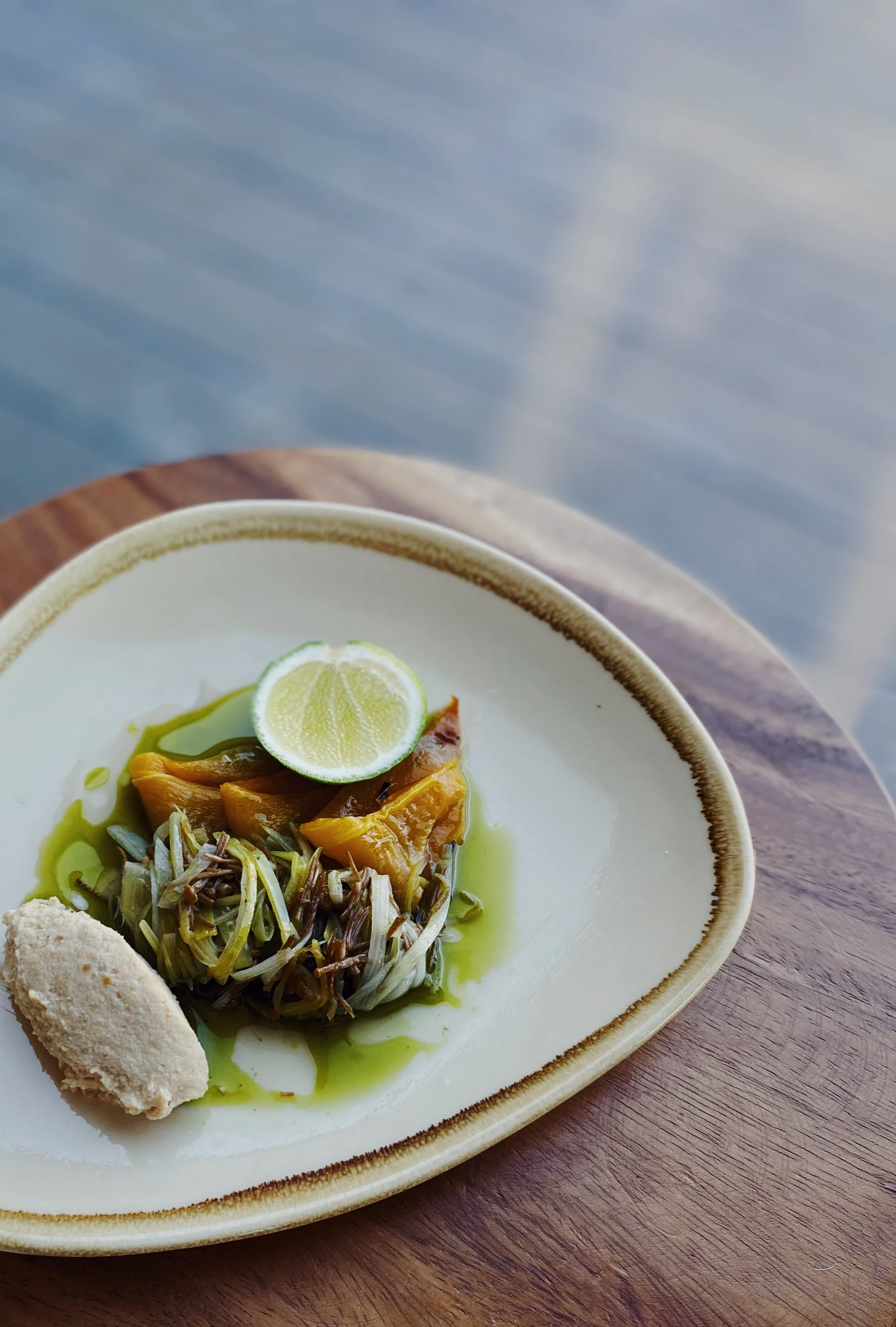 A plate of food with a lime wedge, cooked vegetables, a dollop of paste, and green oil on a white plate with a decorative brown rim, placed on a wooden surface.