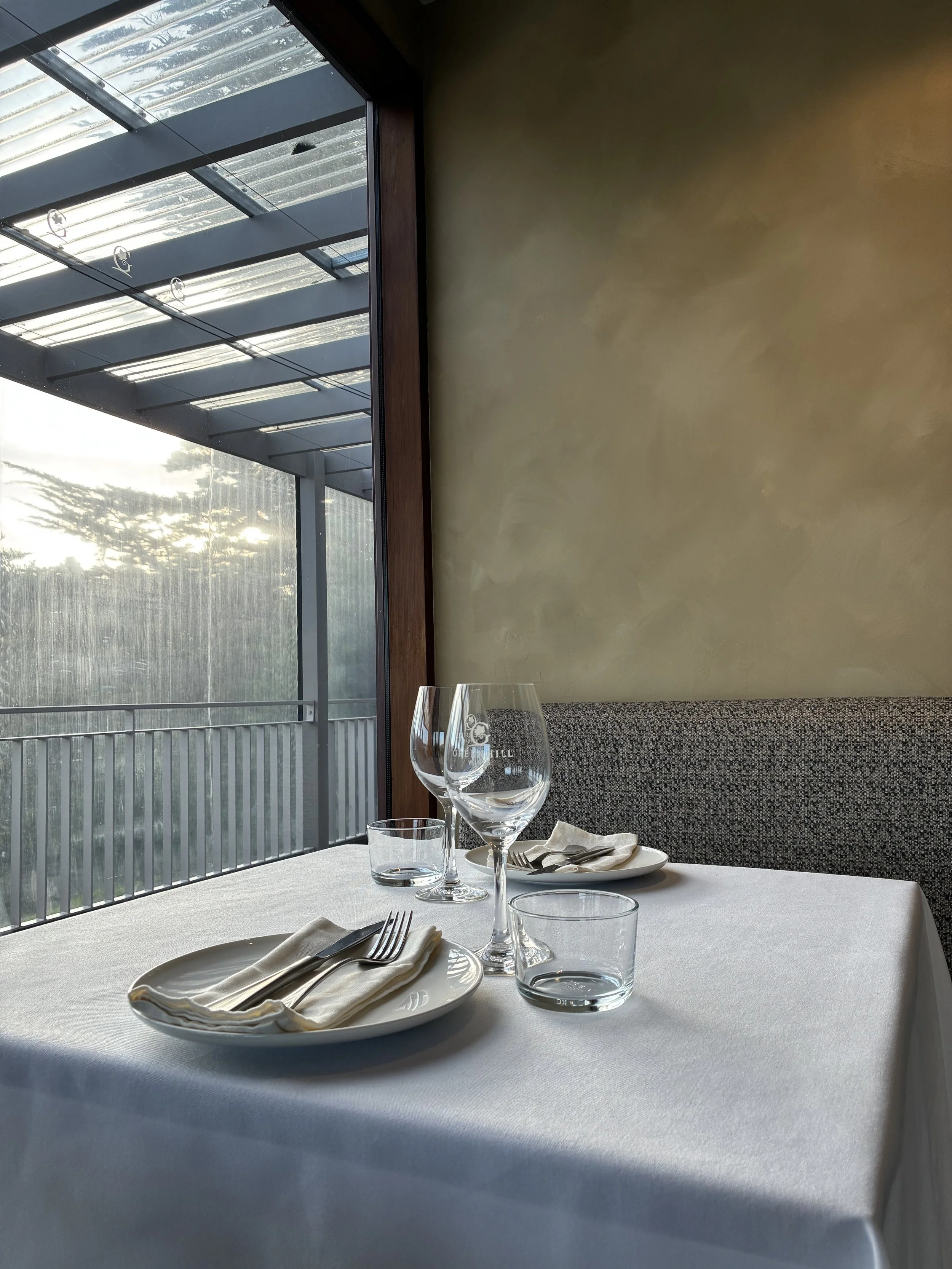 Empty restaurant table set with napkins, forks, wine glasses, a water glass, and a plate, next to a large window showing an outdoor patio with a roof.